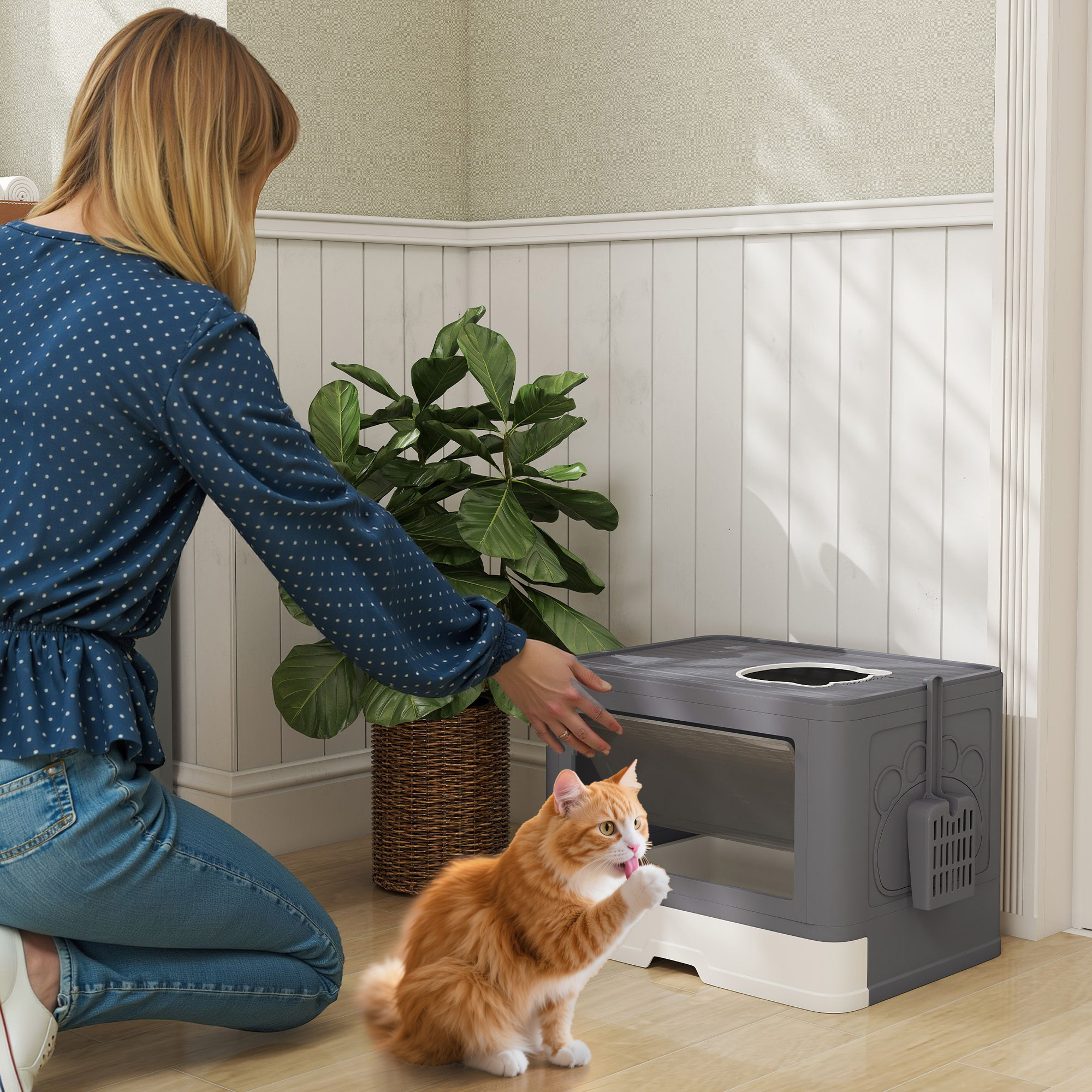 Una mujer se arrodilla junto a una caja de arena mientras un gato naranja se acicala.