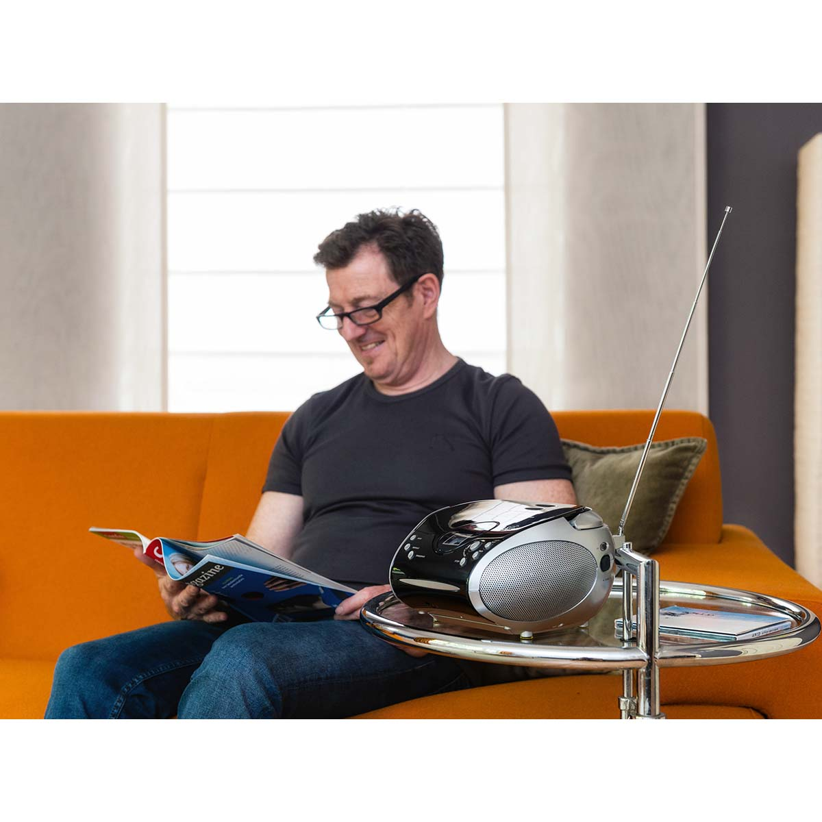A man wearing glasses reads a magazine on an orange couch. A radio sits beside him.