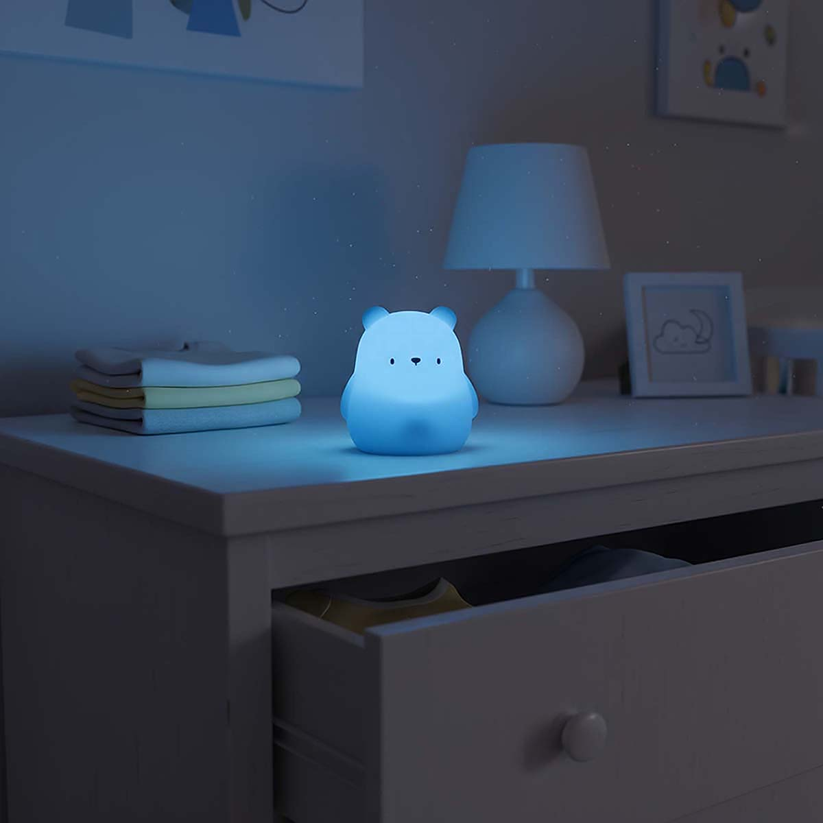Ein beleuchteter Teddybär-Nachtlicht auf einer Kommode neben Kleidung und einem Lampenschirm.
