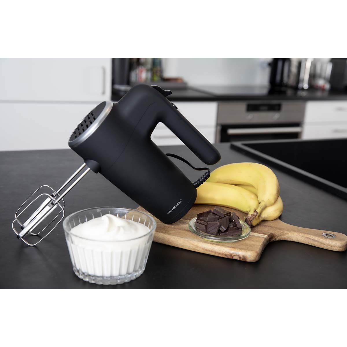 Czarny mikser ręczny z miską śmietany, bananami i kawałkami czekolady na drewnianej desce.