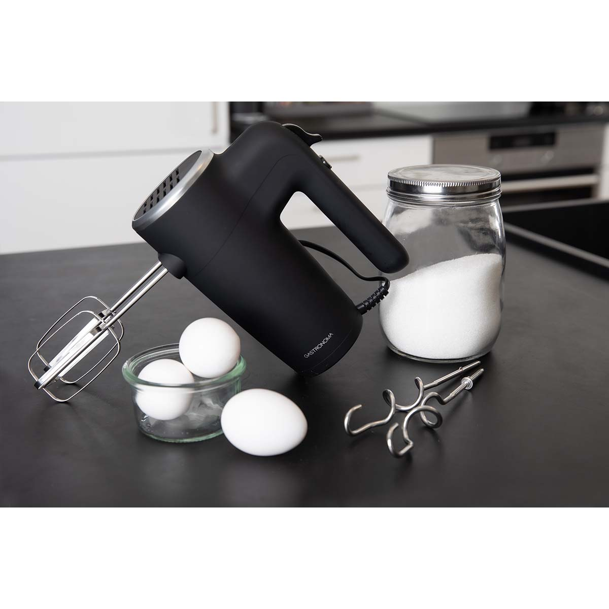 Czarny mikser ręczny z trzepaczkami, jajkami i cukrem w słoiku.