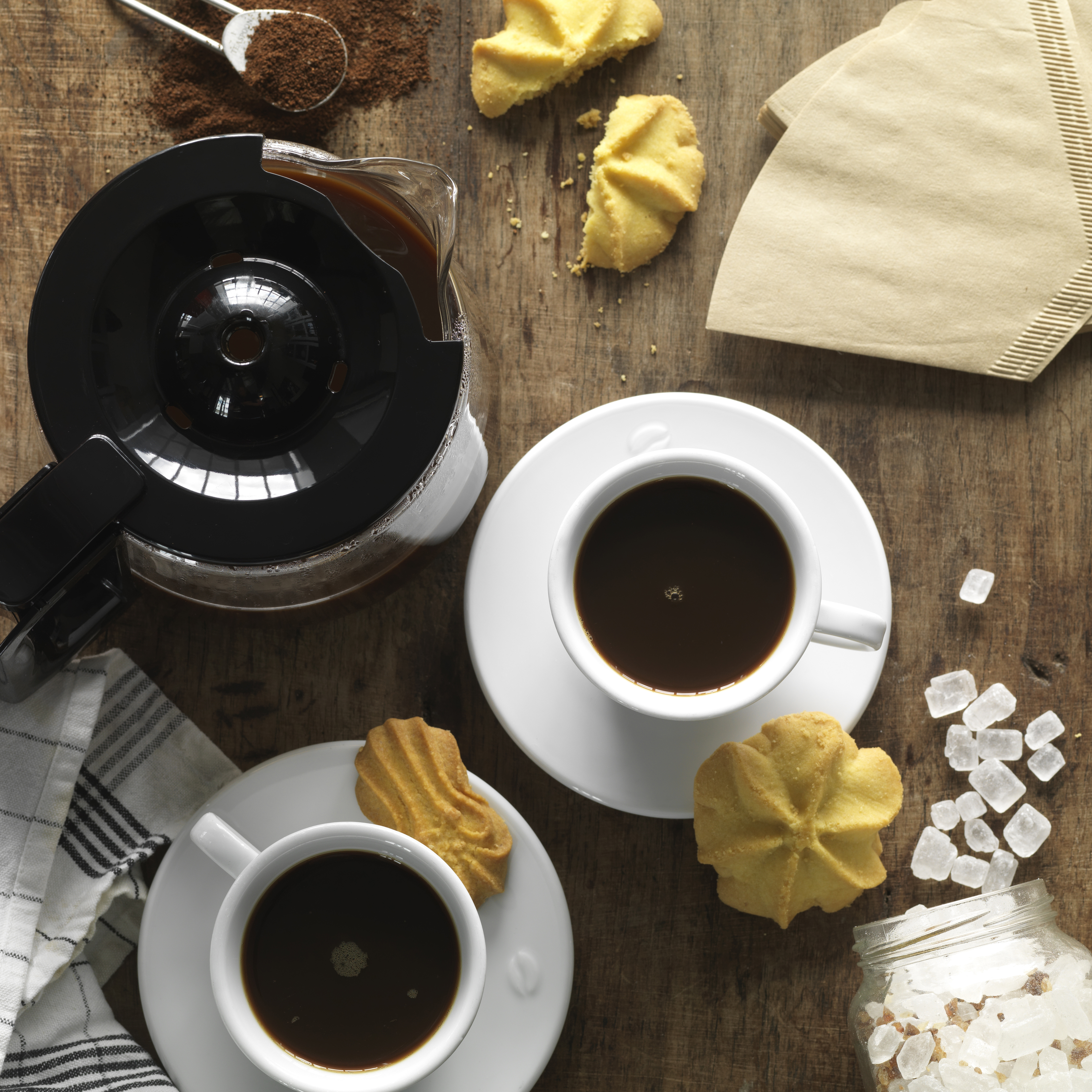 Kaffeekanne, Tassen mit Kaffee, Kekse, Zucker und Filter auf Holztisch.