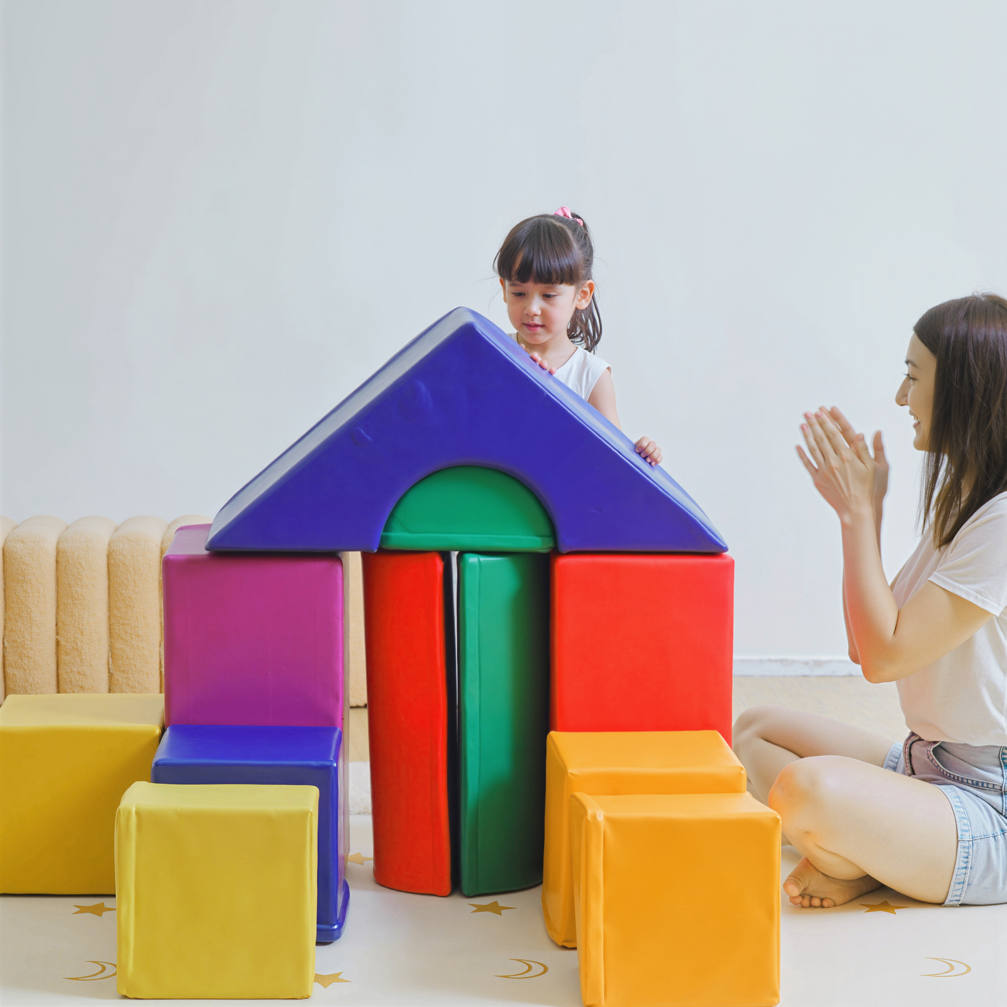 Madre aplaude mientras niña construye casa con bloques.
