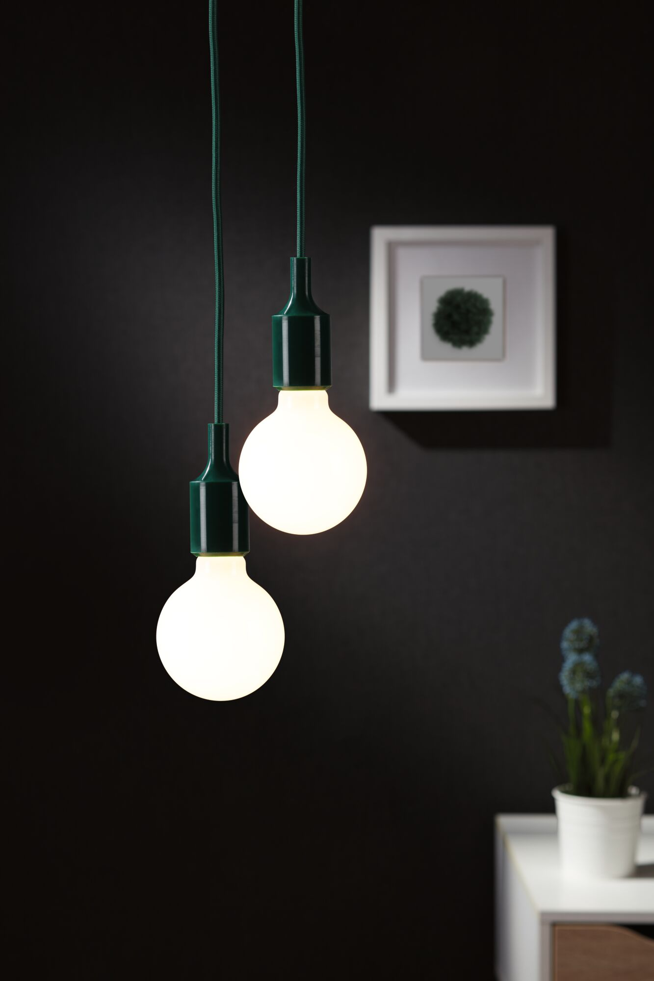 Zwei leuchtende weiße Edison-Glühbirnen hängen an grünen Kabeln vor einer dunklen Wand.