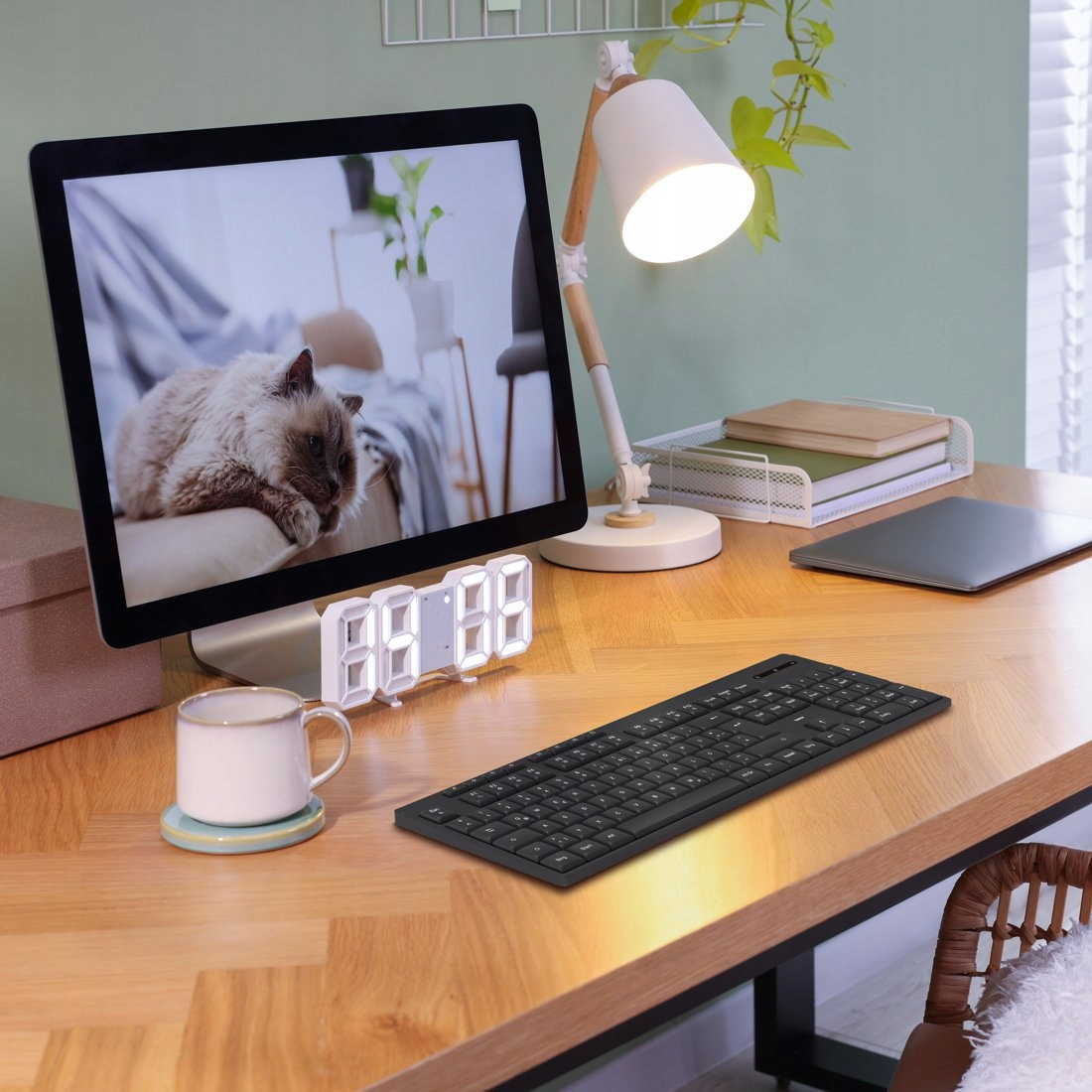 Un escritorio con computadora mostrando un gato, teclado, lámpara, taza y reloj.