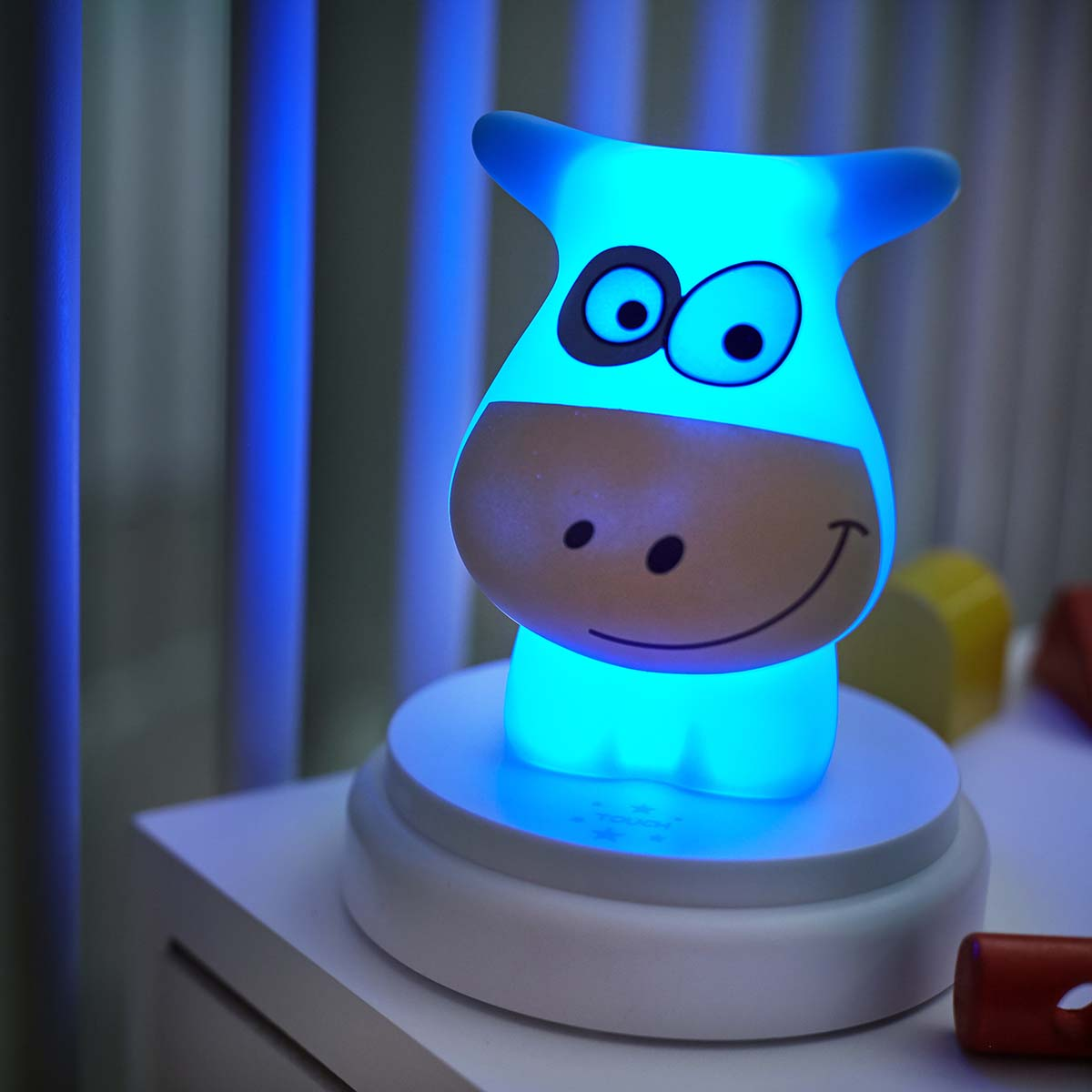 Świecąca lampka w kształcie krowy na stoliku nocnym z niebieskimi światłami w tle.