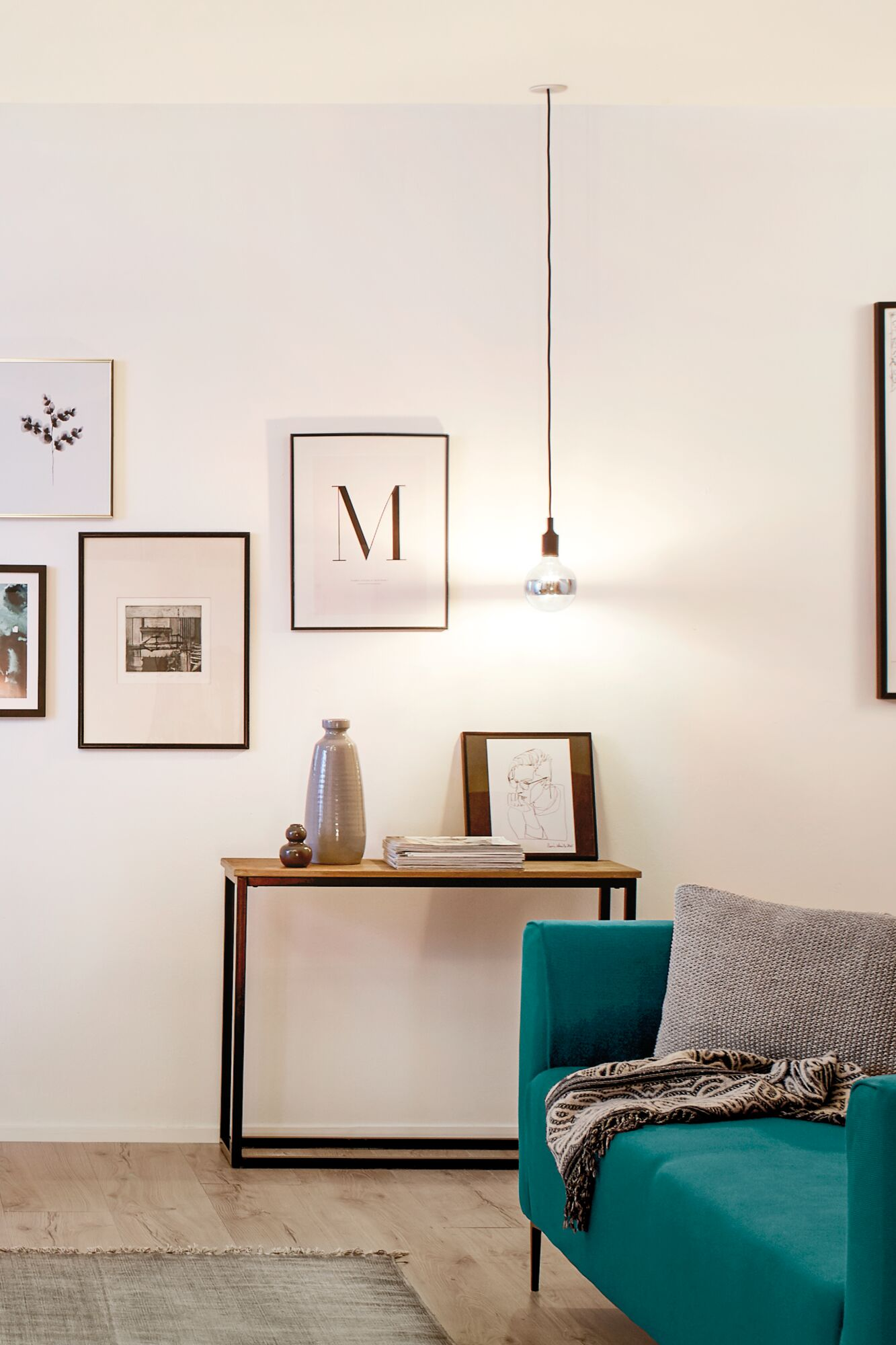 Turkusowy fotel z kocem i poduszką obok konsoli z wazonami i obrazami.