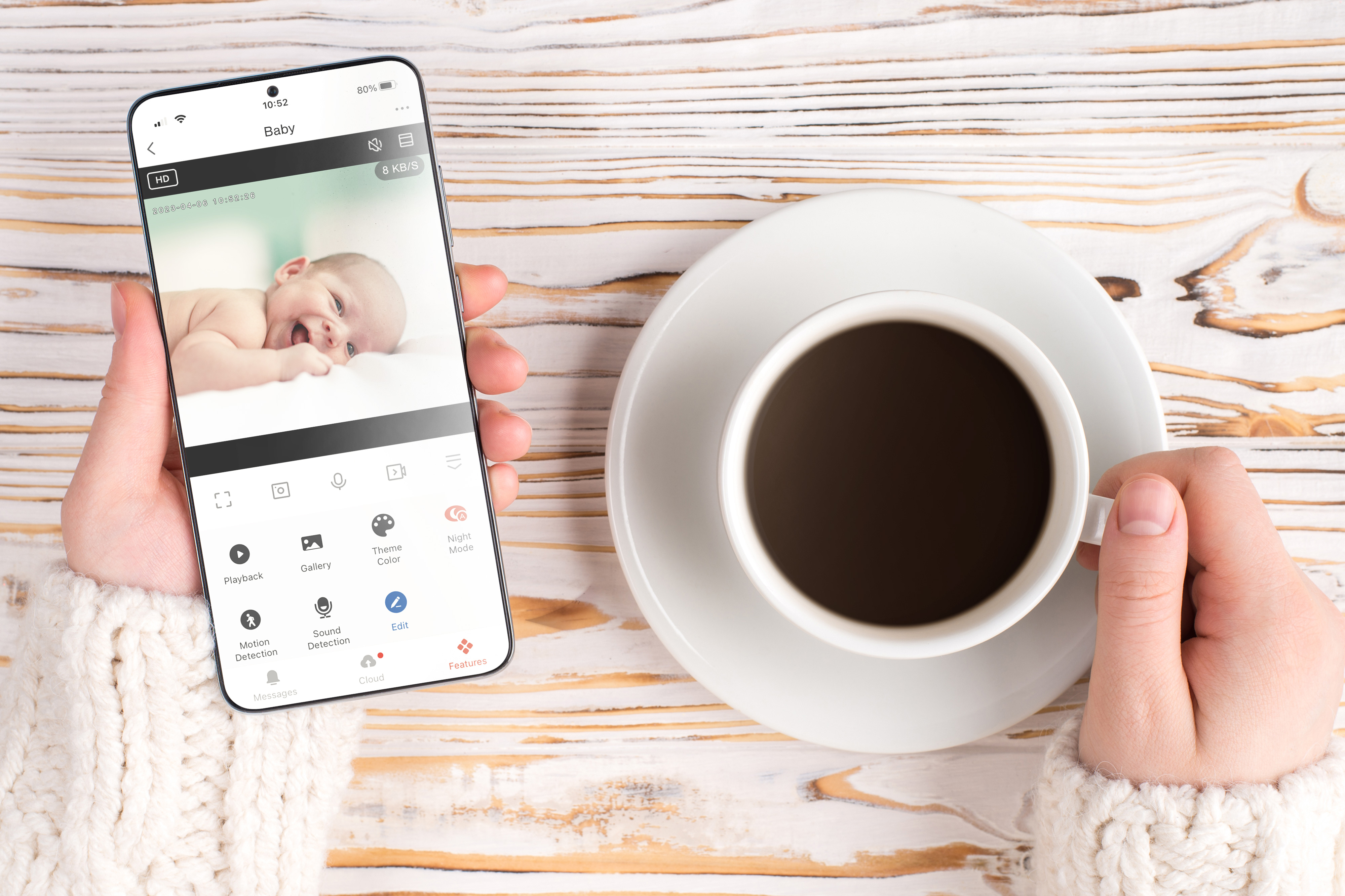 Eine Hand hält ein Handy mit Babyüberwachung. Daneben eine Kaffeetasse.