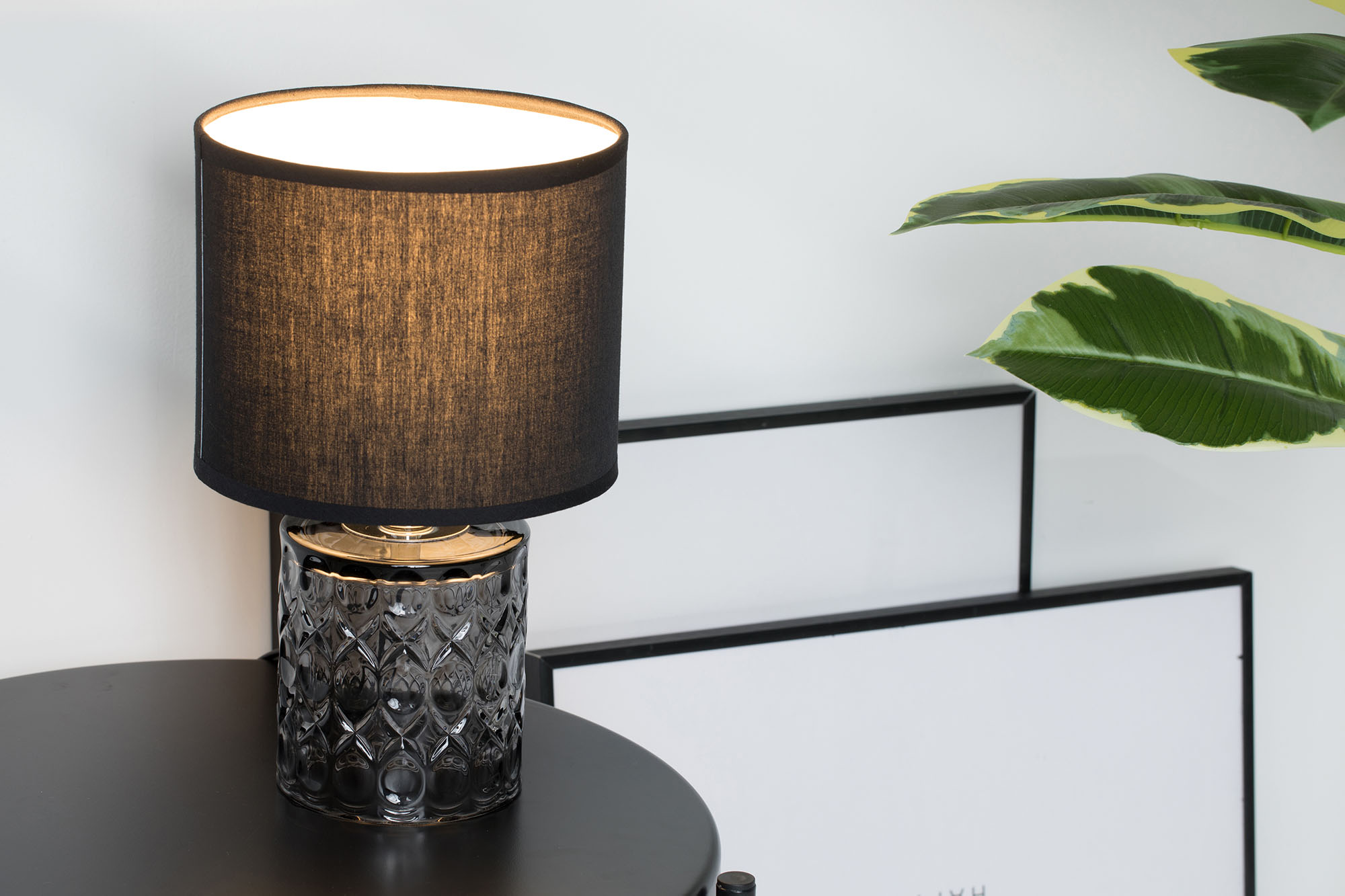 Czarna lampa ze szklaną, wzorzystą podstawą stoi na stole obok rośliny.