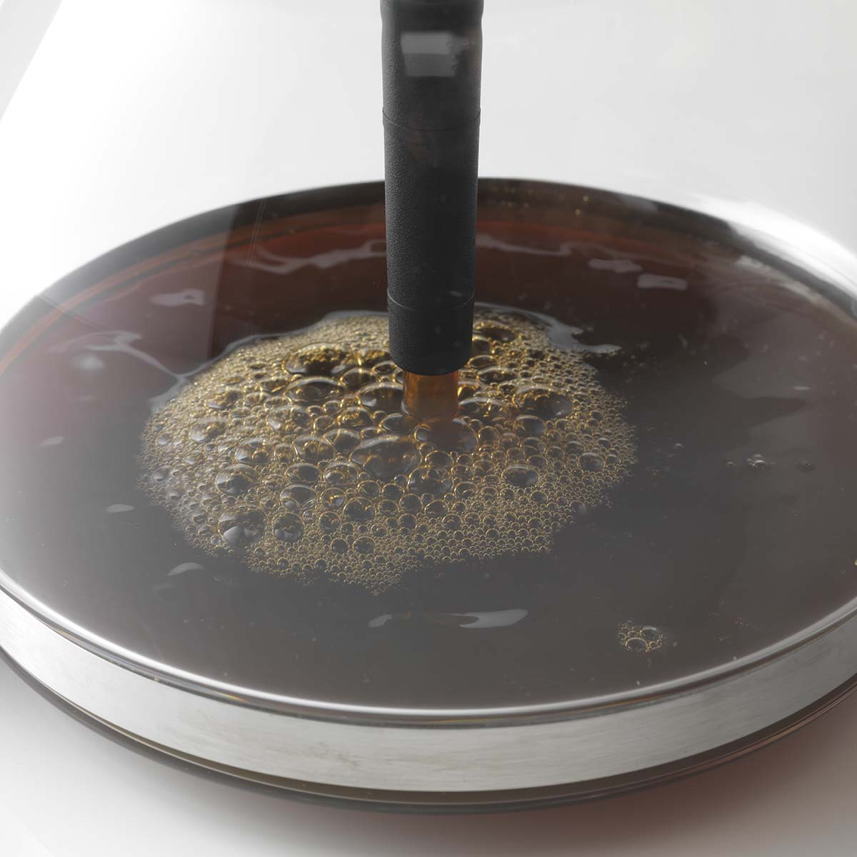 Un'asta nera si immerge in un liquido marrone e schiumoso in un contenitore di vetro con bordo metallico.