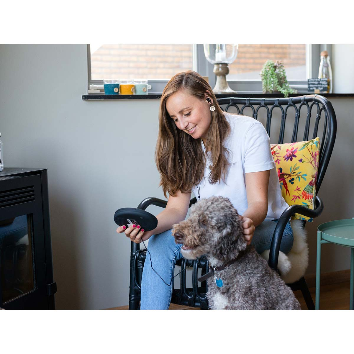 Kobieta siedzi na krześle, trzymając czarne urządzenie i głaszcząc psa.