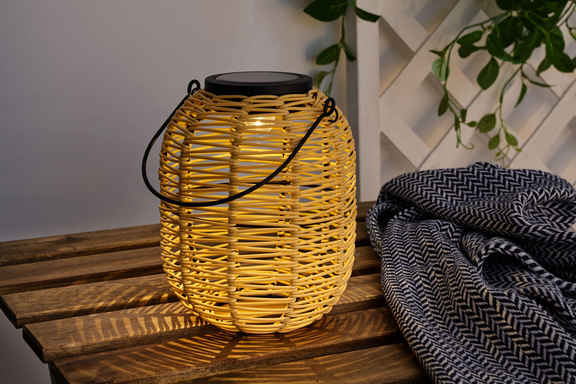 Podświetlona wiklinowa latarnia z czarnym uchwytem stoi na drewnianej ławce obok koca.