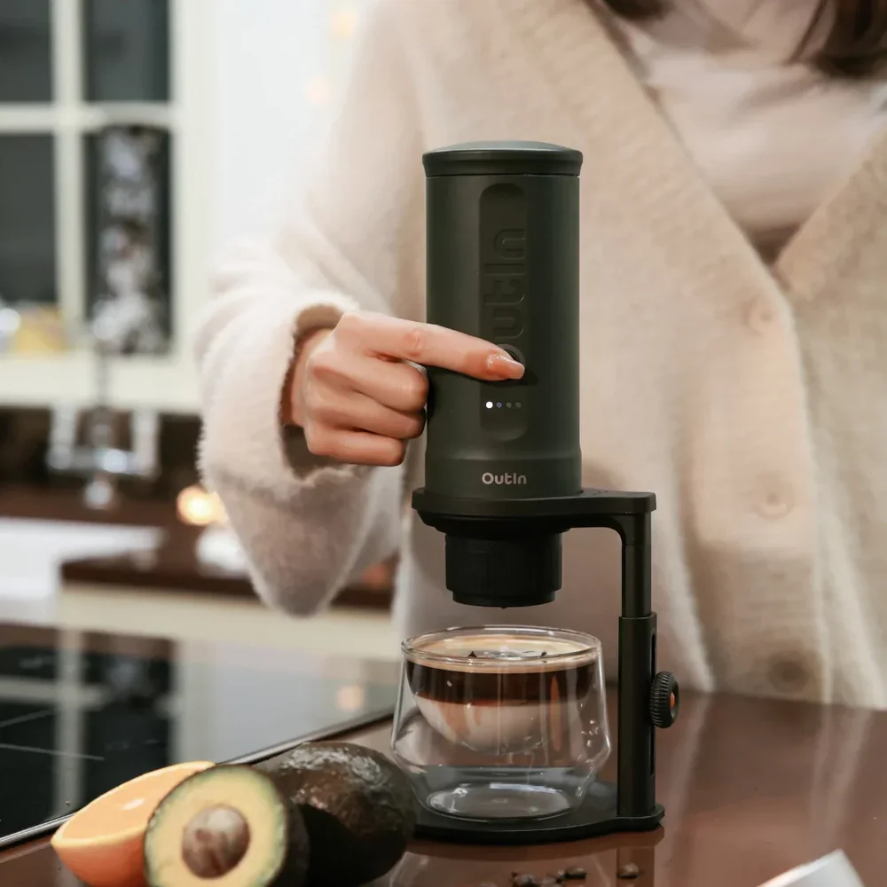 Cafetera verde oscuro operada por una mano, junto a aguacate y rodajas de naranja.