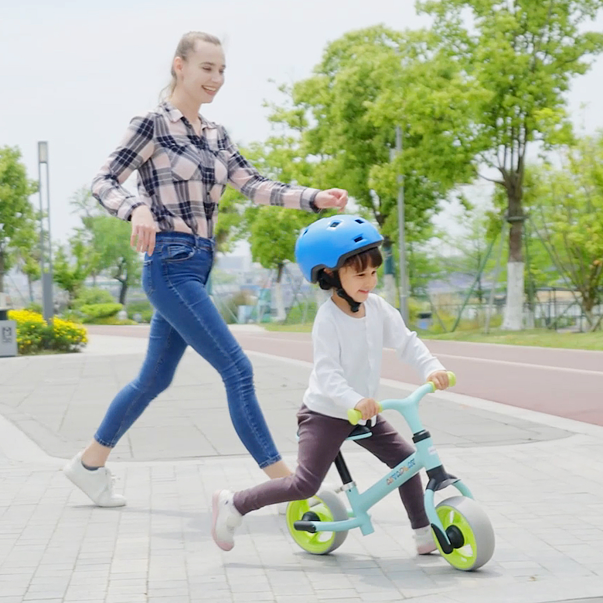 Mujer con camisa a cuadros ayuda a niño con casco azul en bicicleta sin pedales.