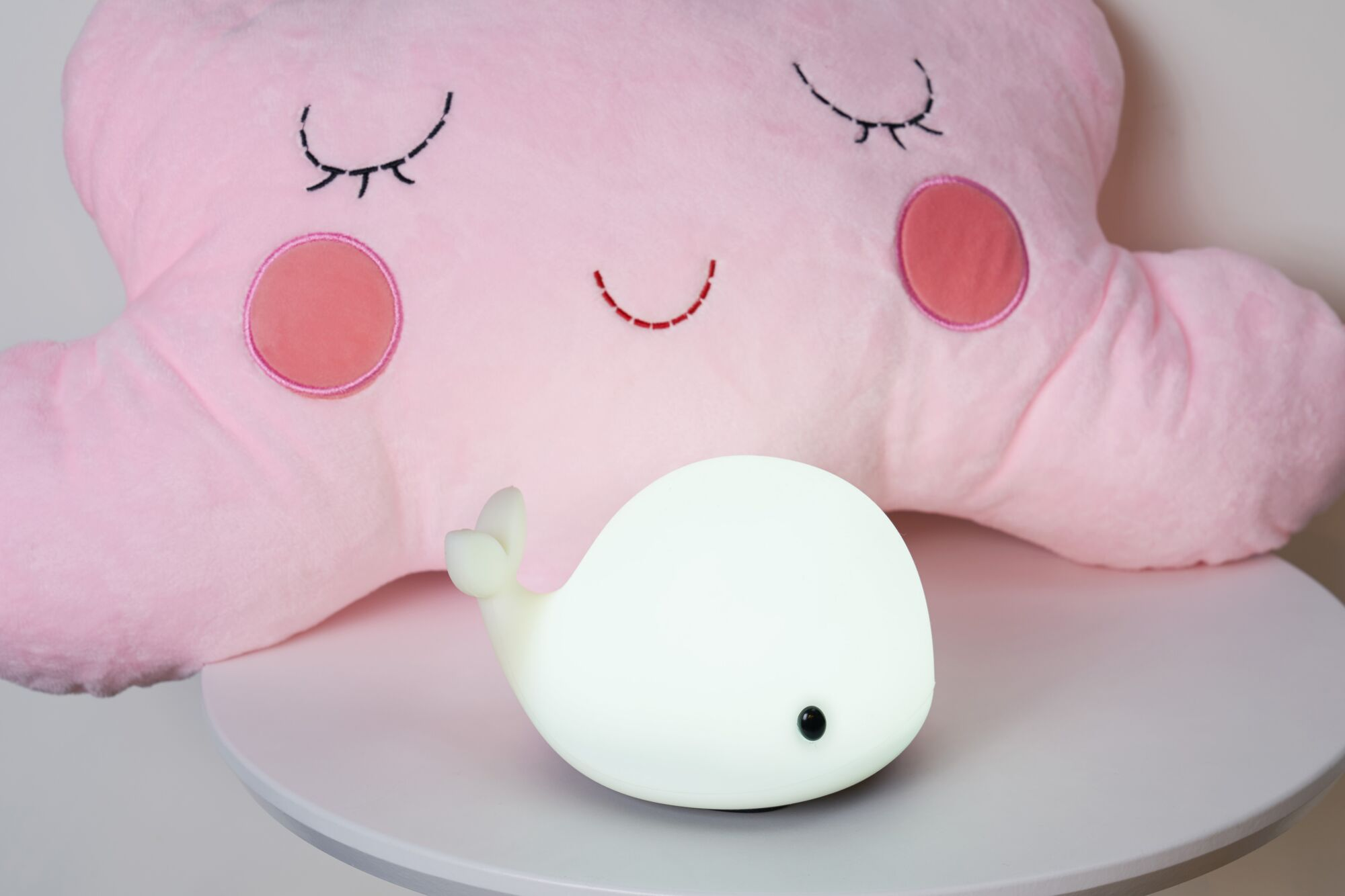 Różowa poduszka w kształcie chmury z naszytymi oczami i policzkami obok świecącej białej lampki wieloryba.