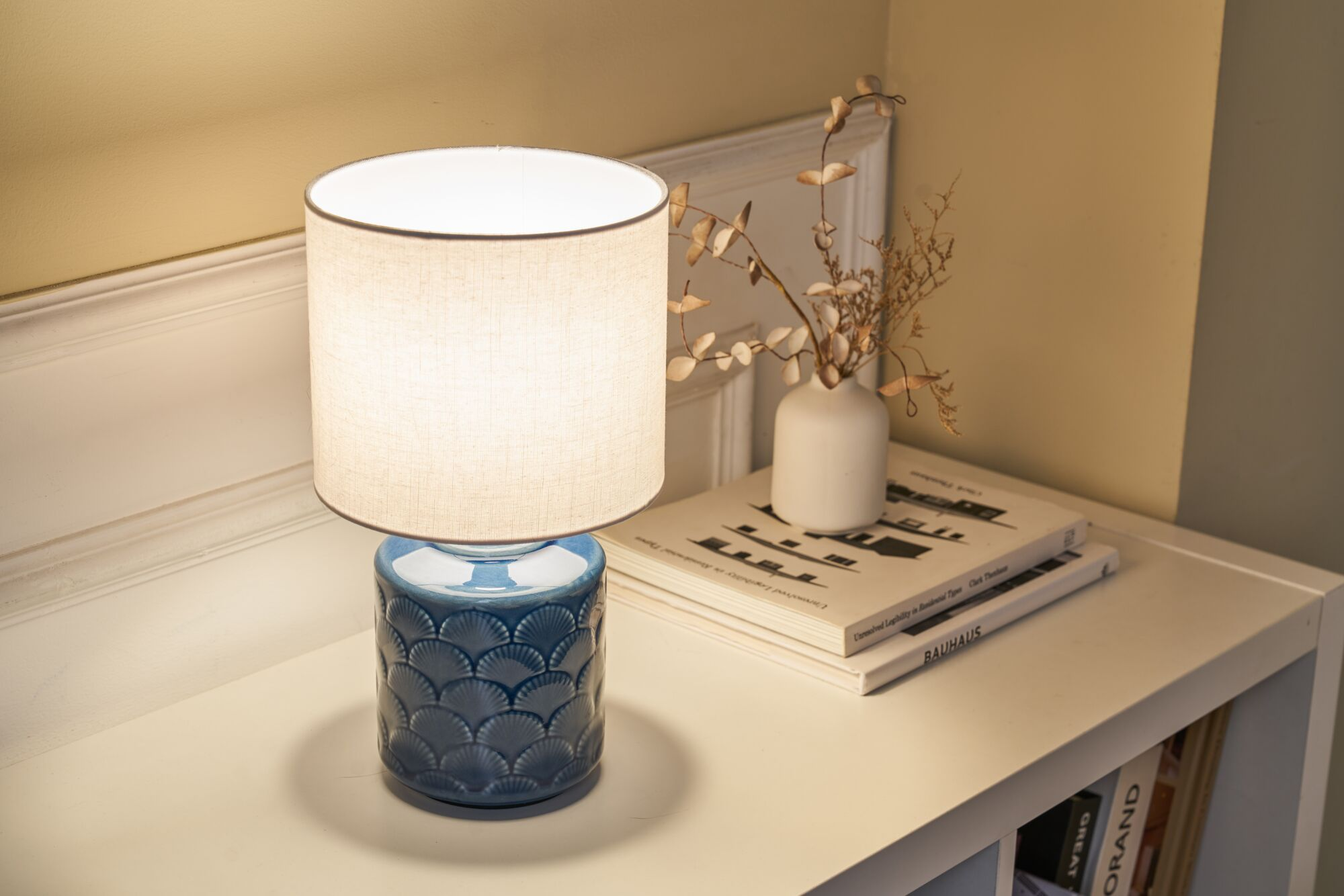 Niebieska lampa z reliefem muszli na stole obok książek i wazonu z suszonymi kwiatami.
