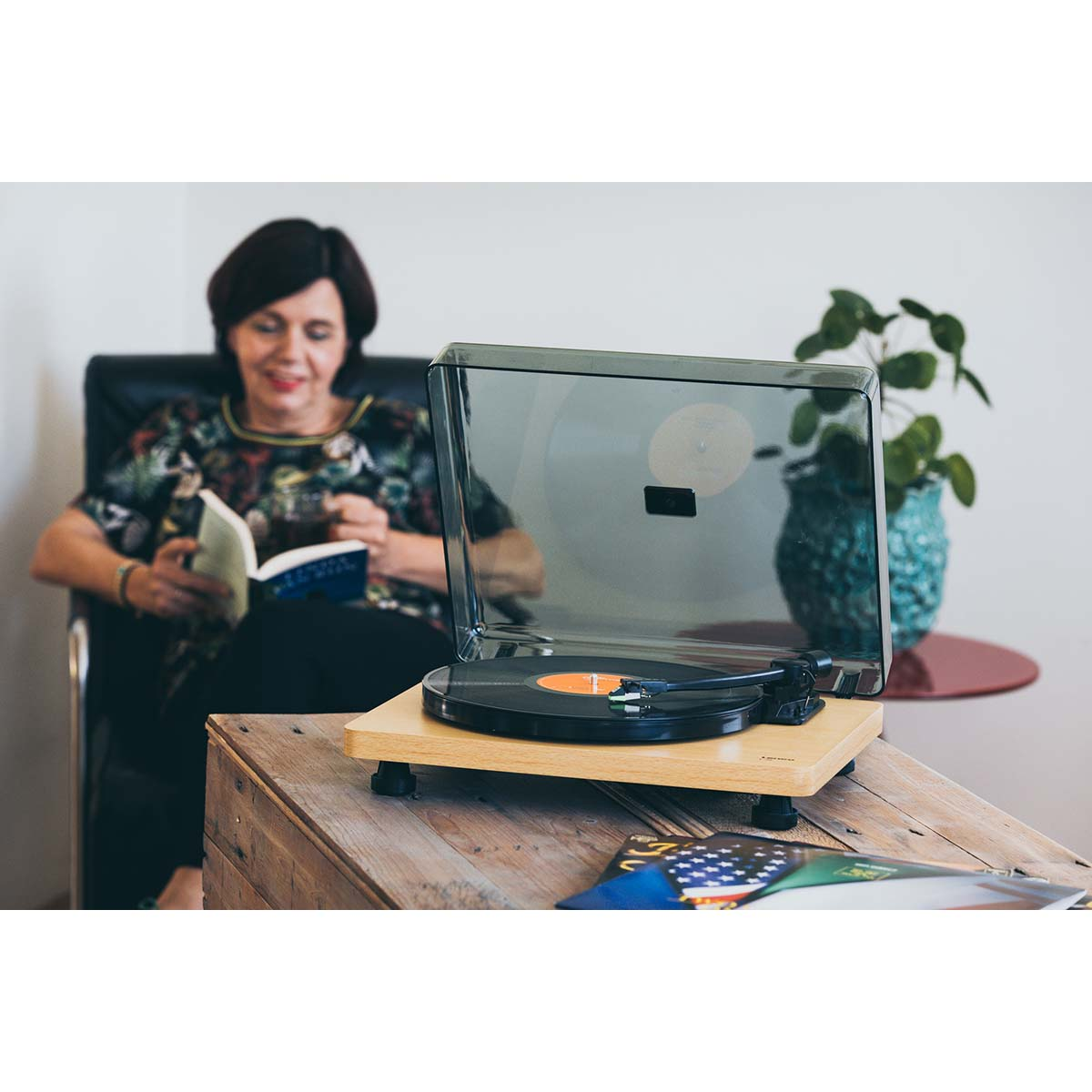 Kobieta czyta książkę, gramofon z płytą, roślina w doniczce z tyłu.