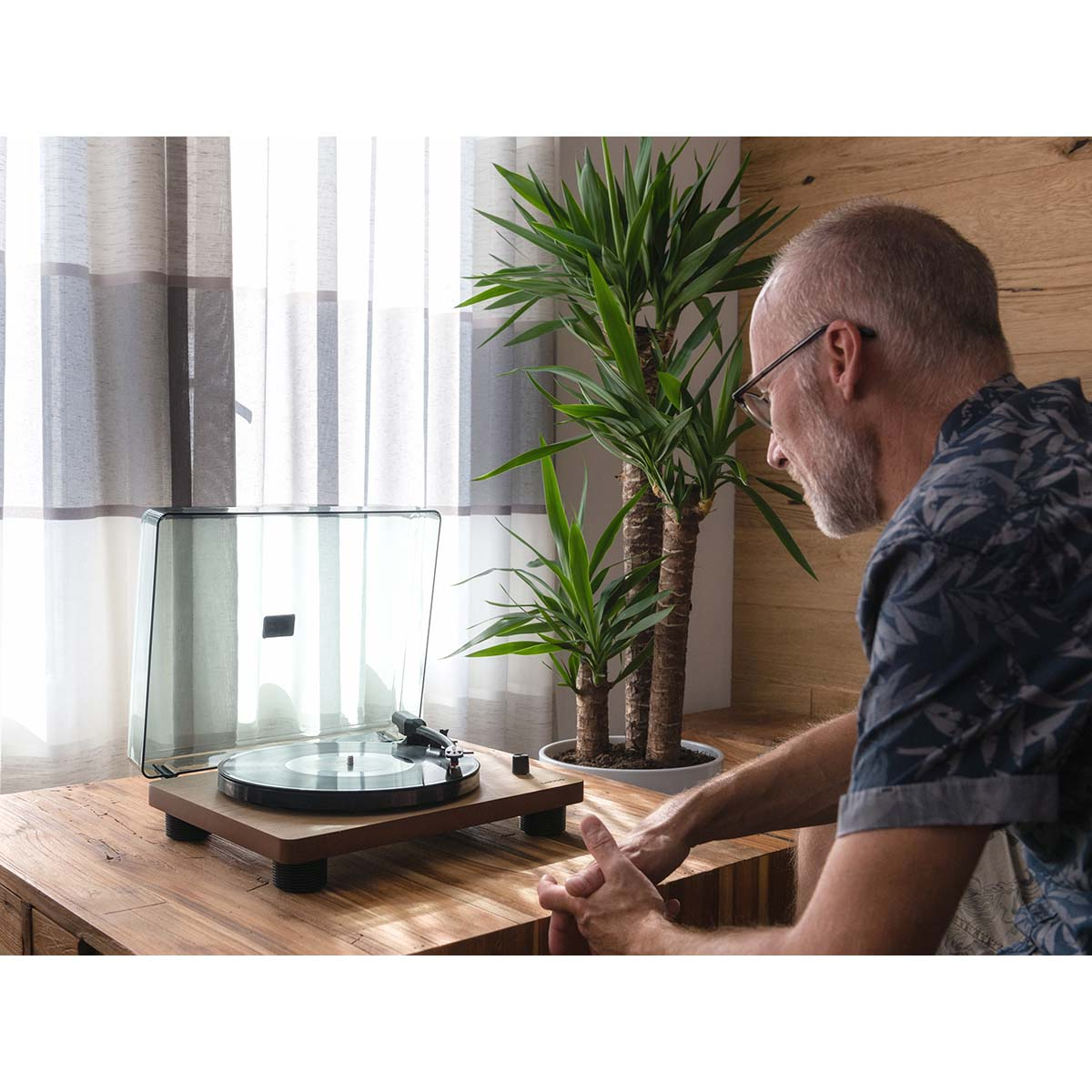 Mężczyzna siedzi przed gramofonem z płytą winylową i dwoma roślinami juki.