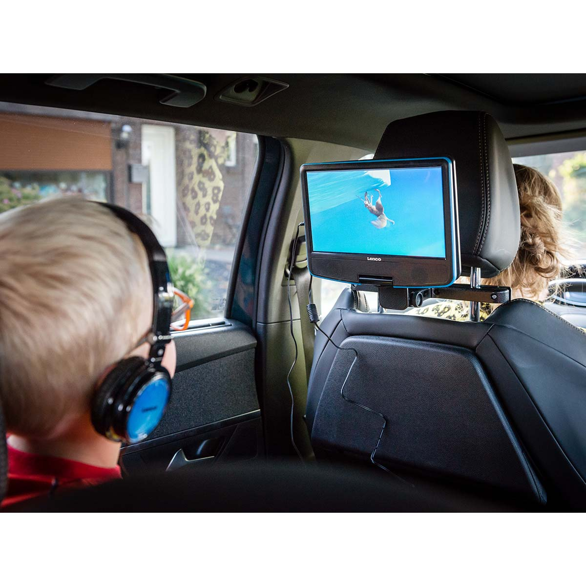 Dziecko w słuchawkach w samochodzie ogląda ekran zamontowany na oparciu. Widoczna scena zewnętrzna.