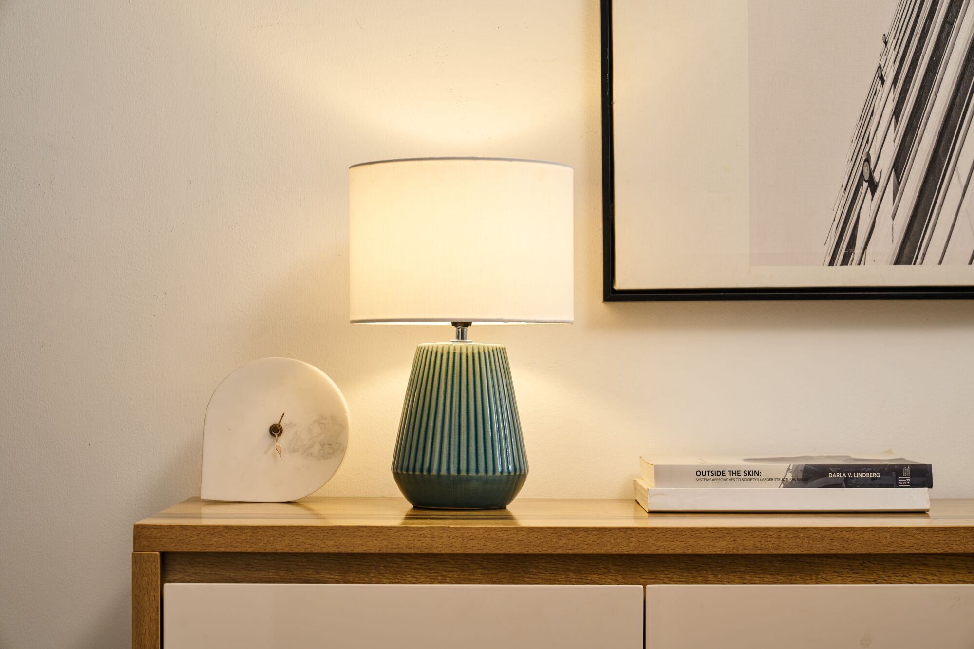 Zielona podstawa lampy z białym kloszem, obok książki i rzeźby.