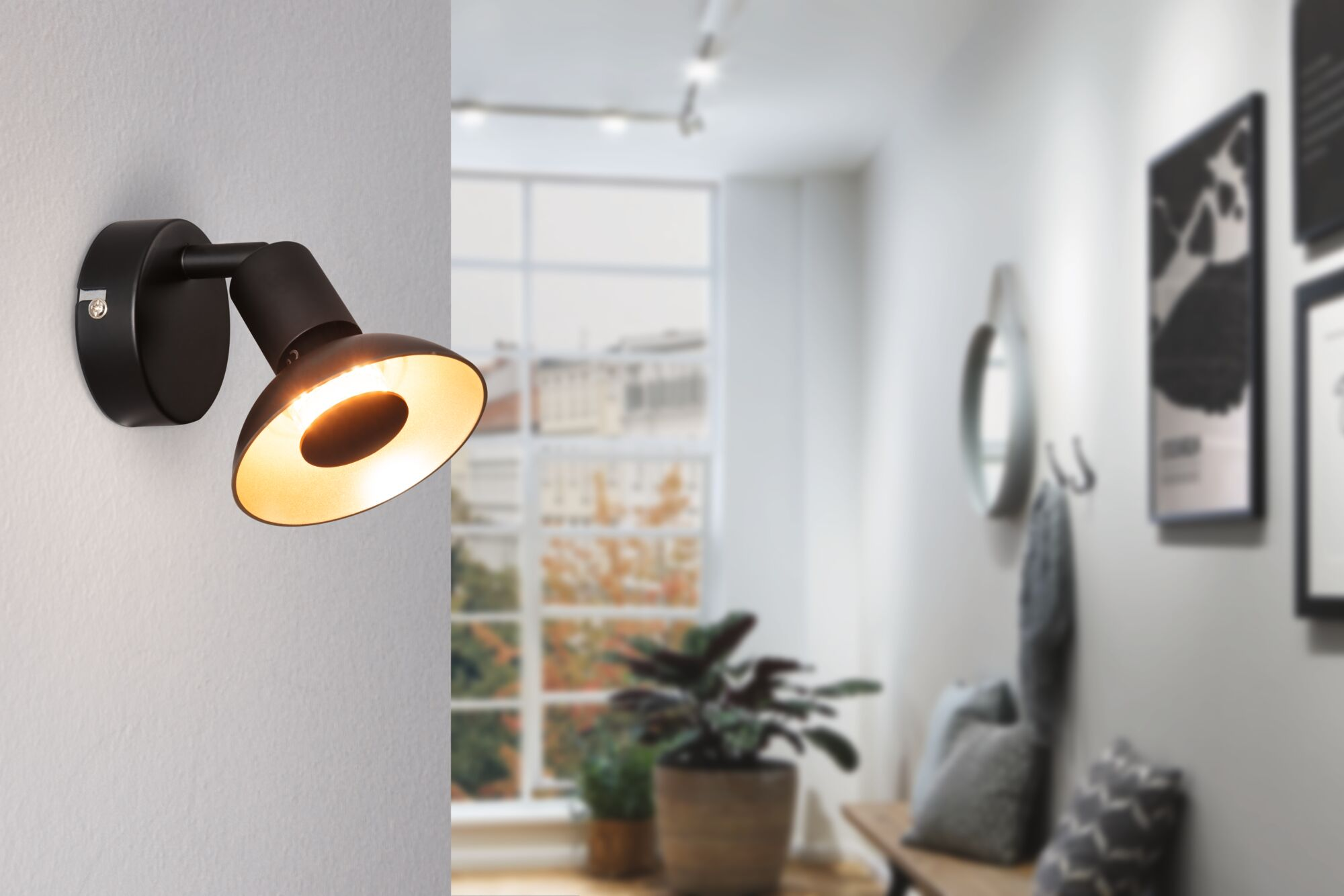 Ein schwarzer Wandstrahler leuchtet warmweiß auf einer weißen Wand. Im Hintergrund ein Fenster.