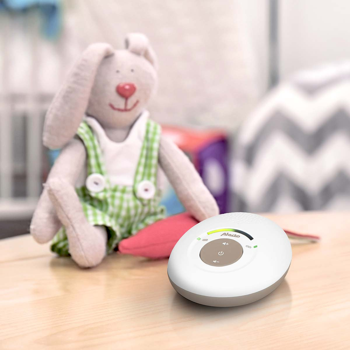 A stuffed bunny in overalls sits next to a white baby monitor on a wooden table.