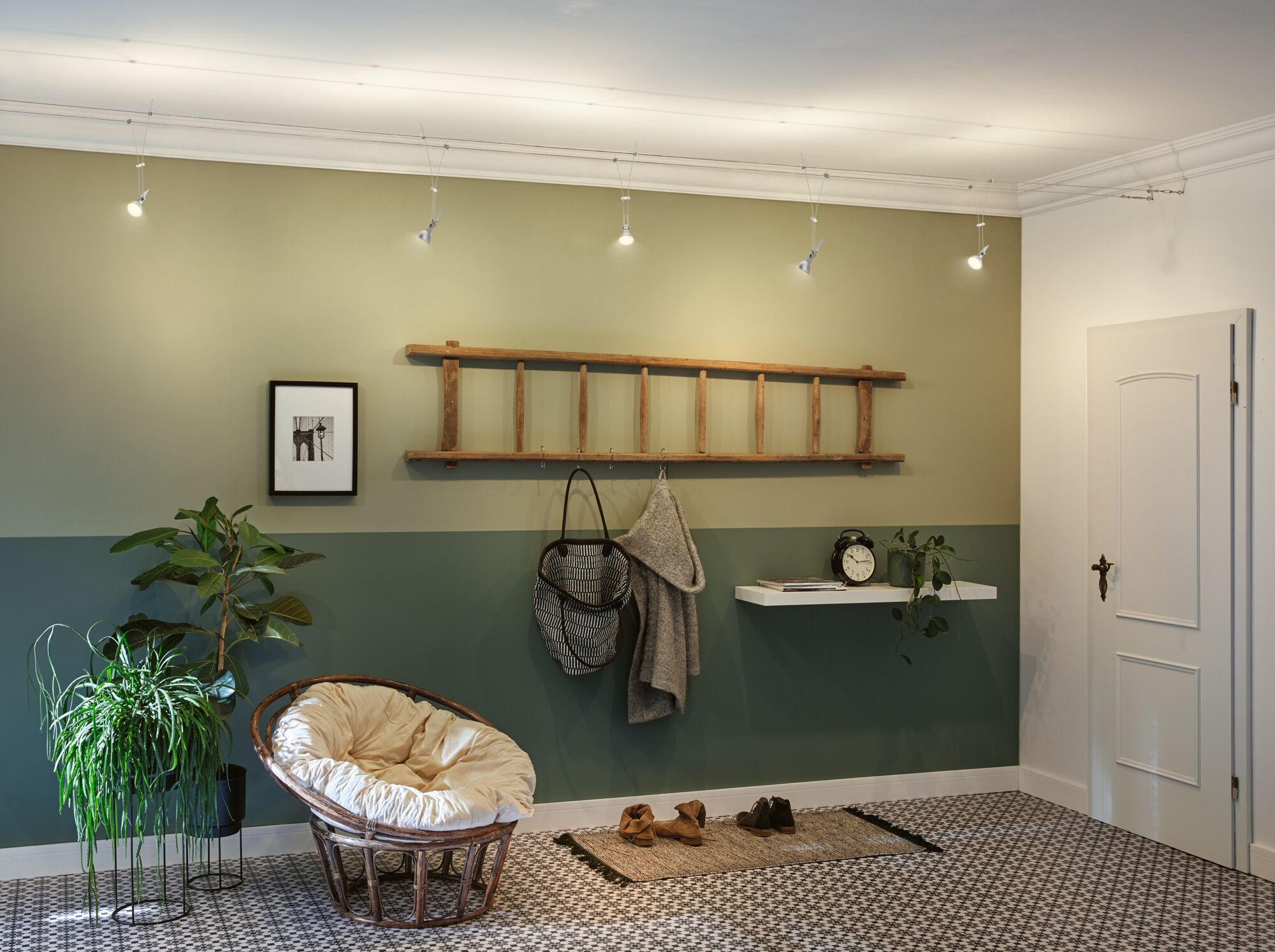 Un corridoio con muro verde, poltrona in rattan, piante, scala, porta e pavimento a motivi.
