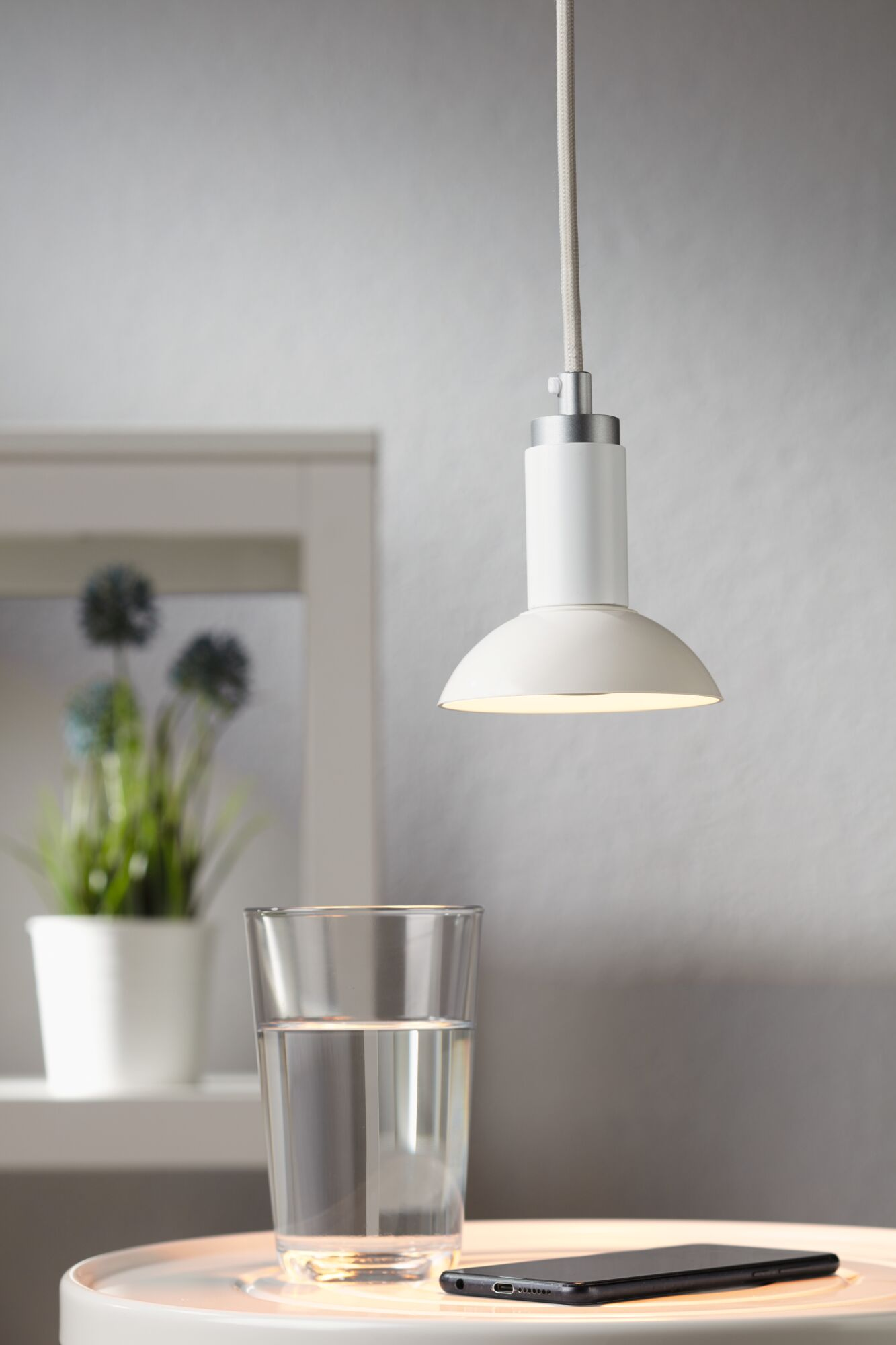 Biała lampa wisząca wisi nad stołem ze szklanką wody i smartfonem.