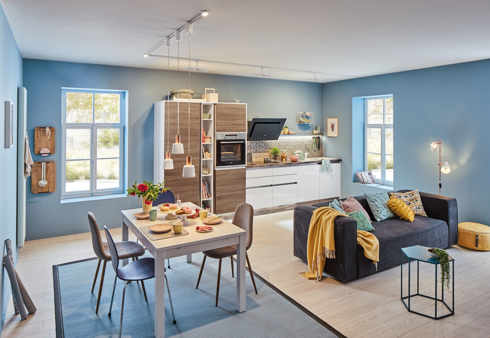 Ein Wohnzimmer mit Essbereich und Küche in Blautönen, heller Einrichtung und Holzakzenten.