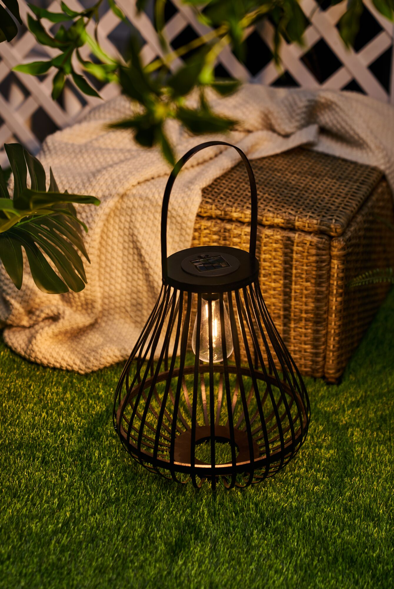 Czarna latarnia z żarówką stoi na zielonej trawie obok koca i kosza.