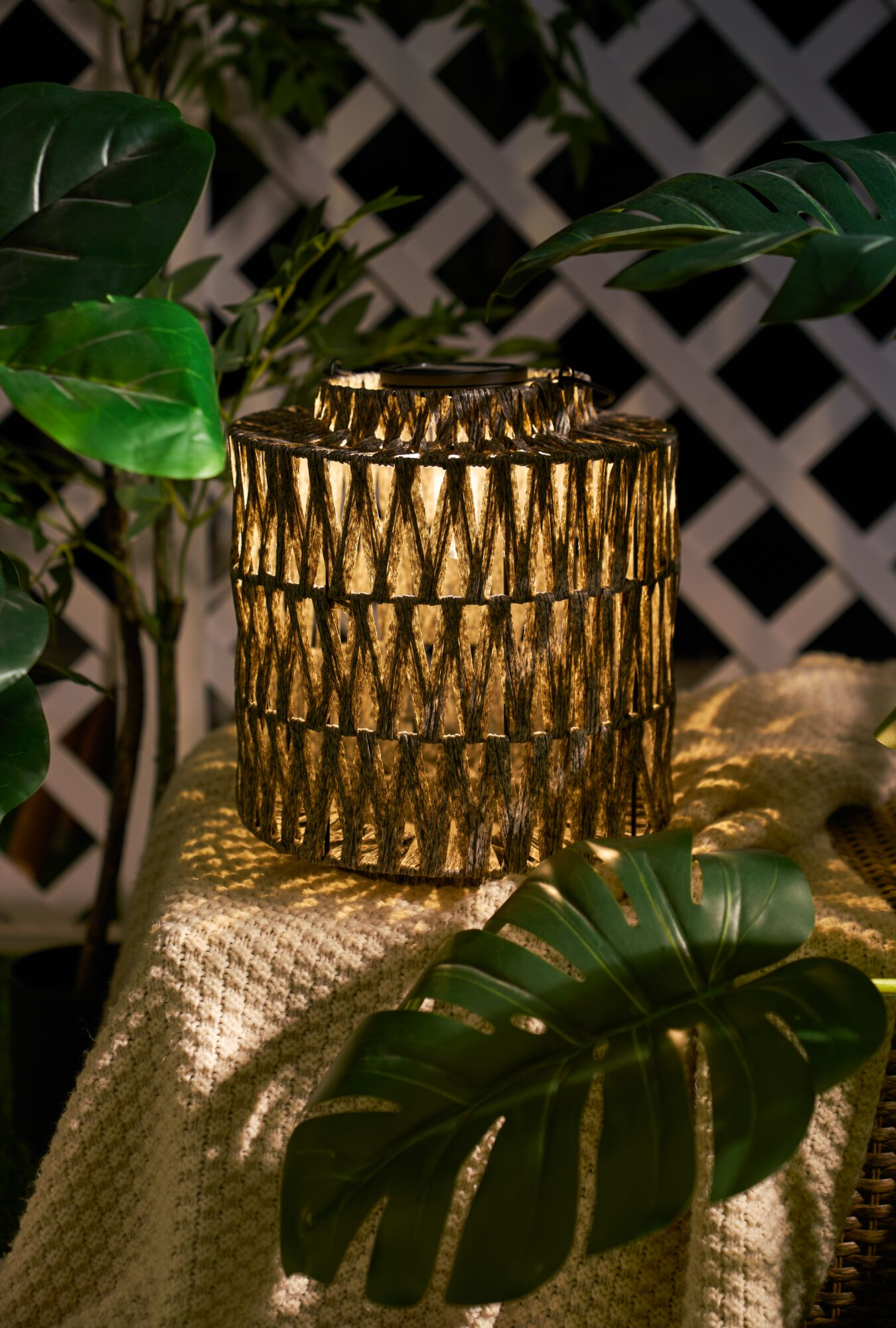 Podświetlona latarnia z plecionym wzorem, otoczona zielonymi liśćmi i kocami.