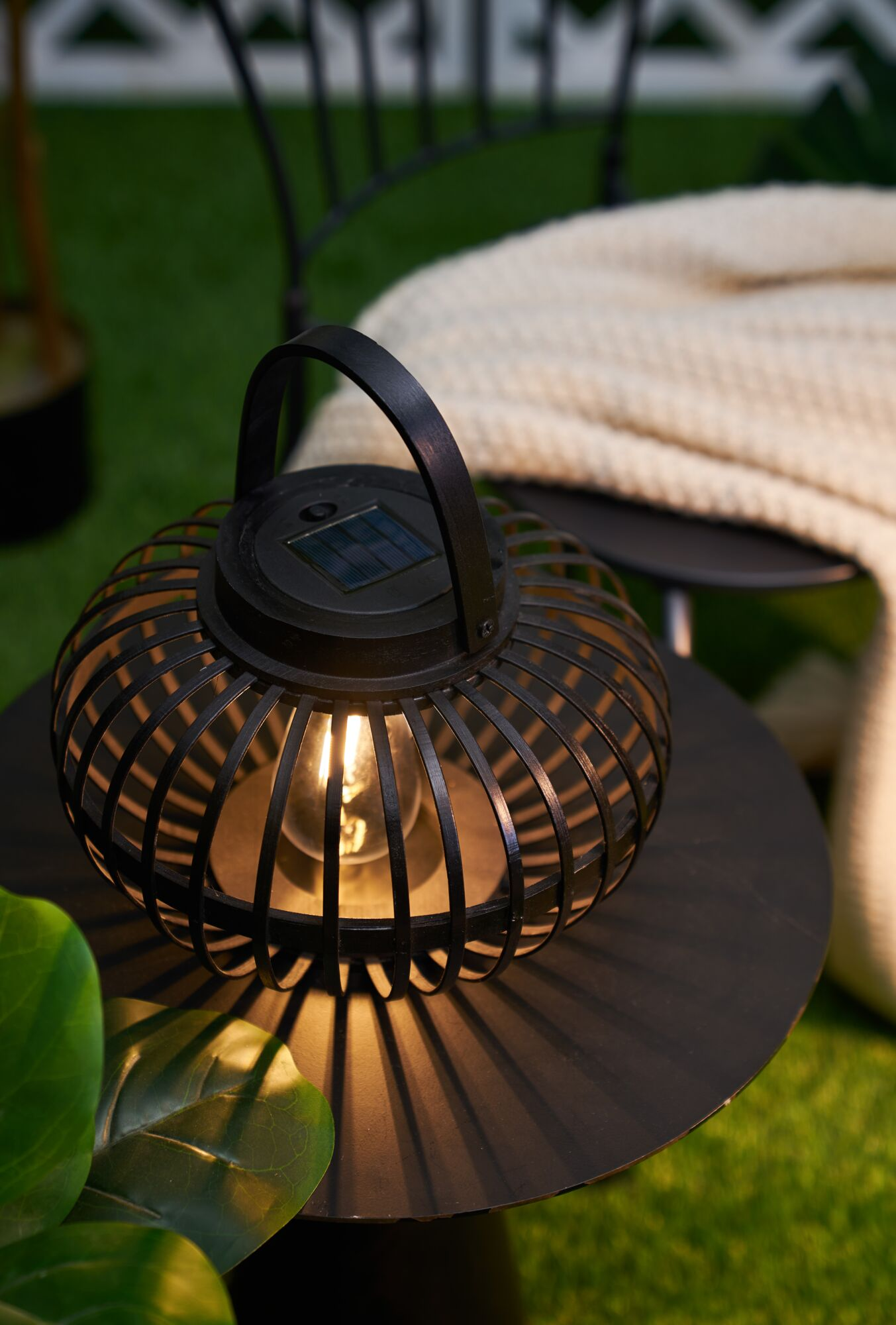 Czarny stolik z latarnią, podświetloną żarówką i cieniami na trawie.