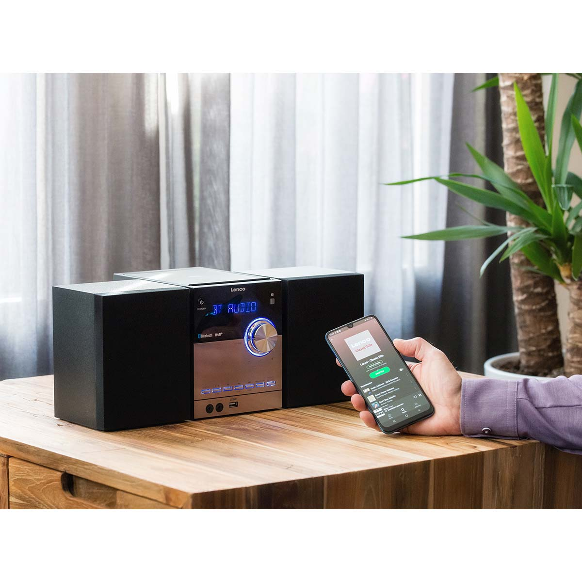 Ein Mann hält ein Smartphone, das auf einen Lenco-CD-Player mit Lautsprechern zeigt.