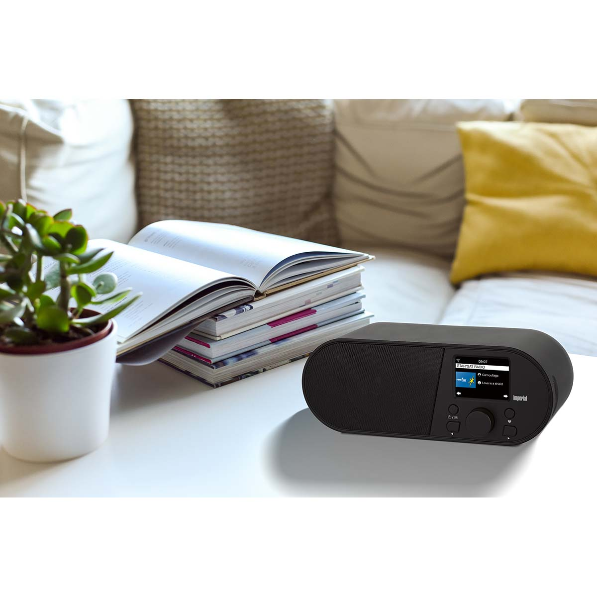 A black digital radio sits on a table next to open books and a potted plant.