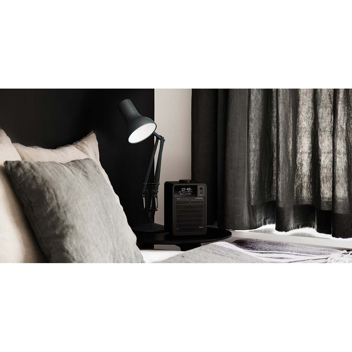 A black desk lamp shines on a bedside table with a radio and pillows.