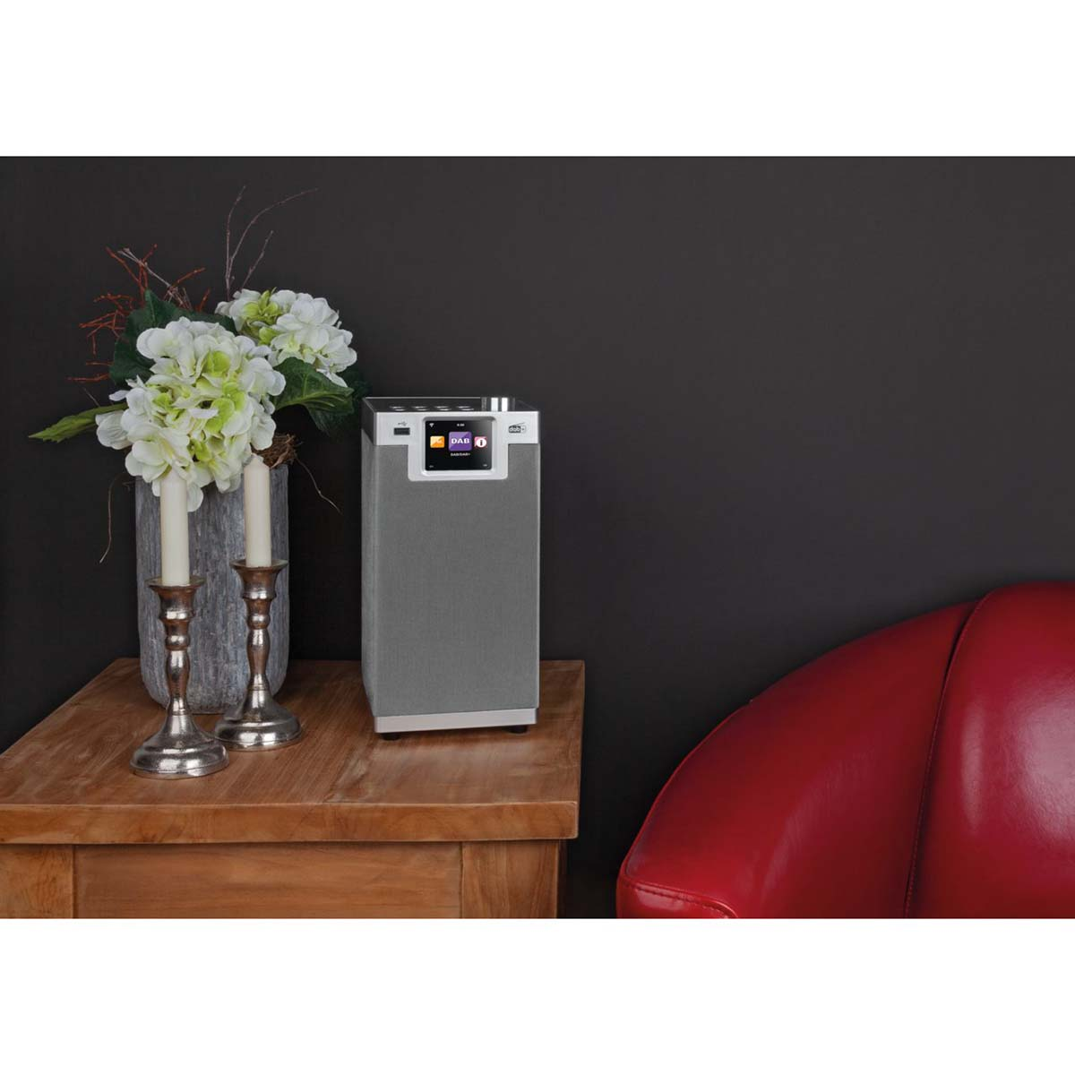 Gray DAB radio with silver display on wooden table, next to candles and flowers.