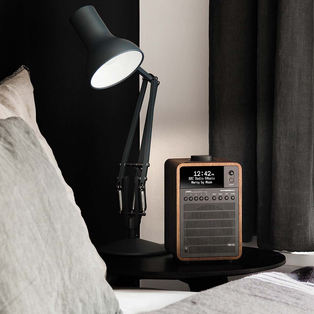 Une lampe de bureau sombre éclaire une radio en bois sur une petite table noire près d'un lit.