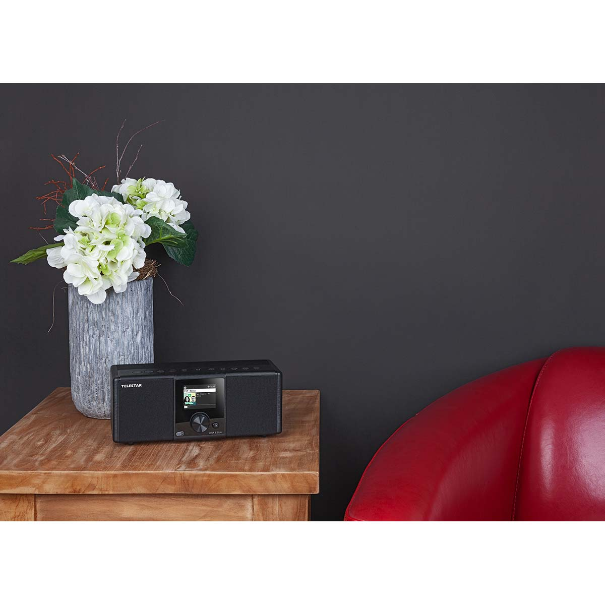 Black radio on wooden table next to flowers and red armchair.