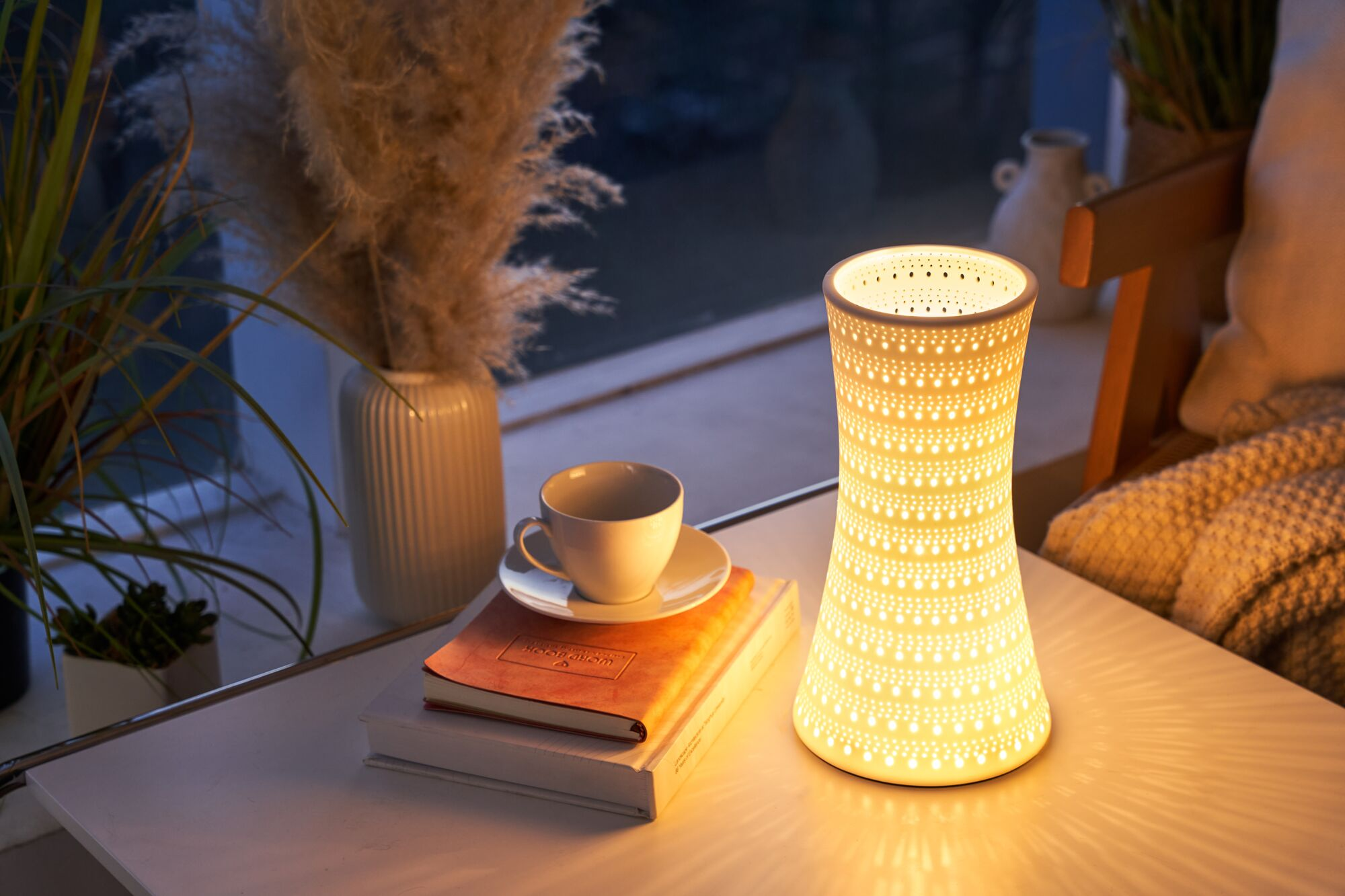 Podświetlana lampa stoi na stole obok książek, filiżanki i roślin.