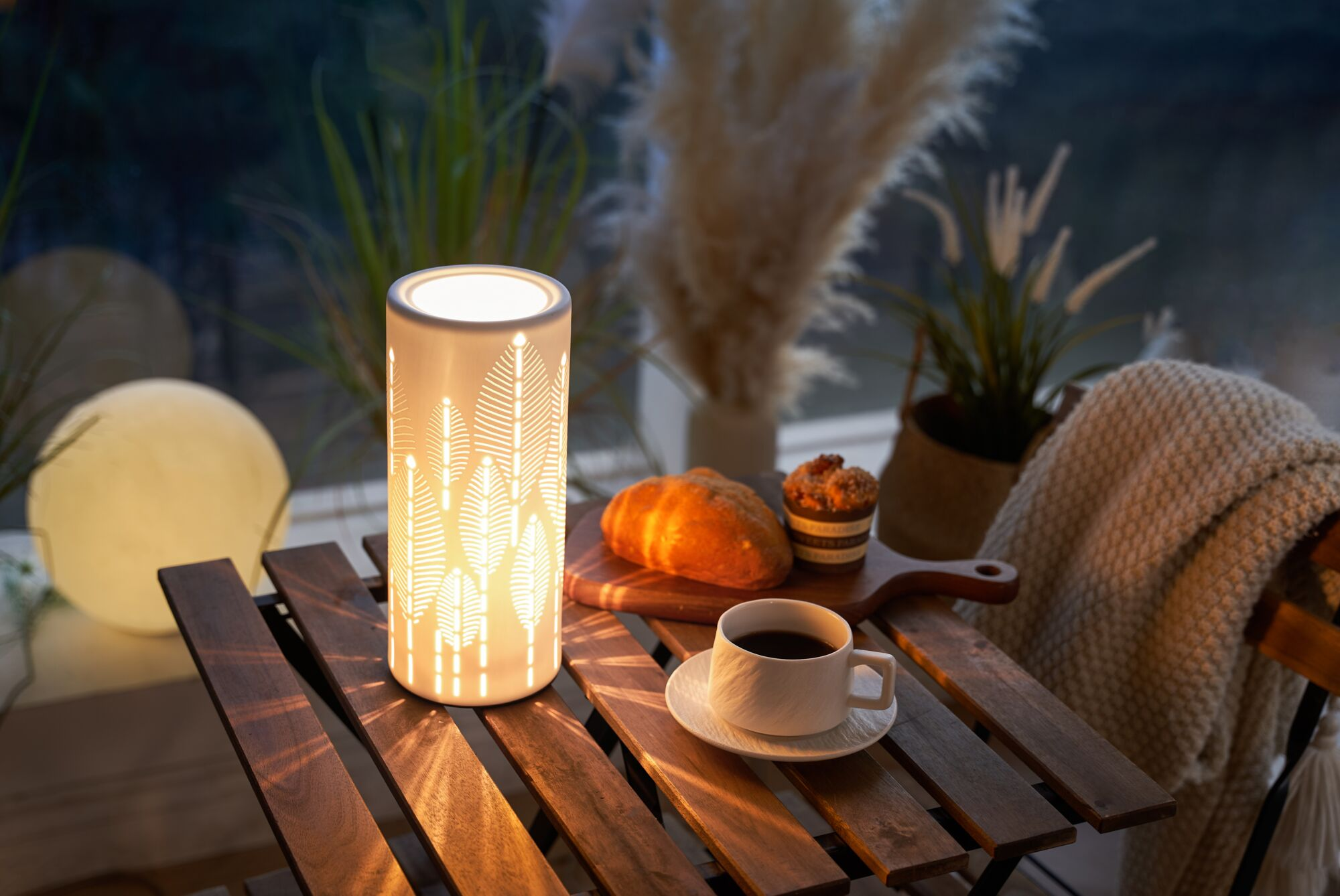 Podświetlona latarnia ze wzorem liści stoi na drewnianym stole z kawą i ciastkami.