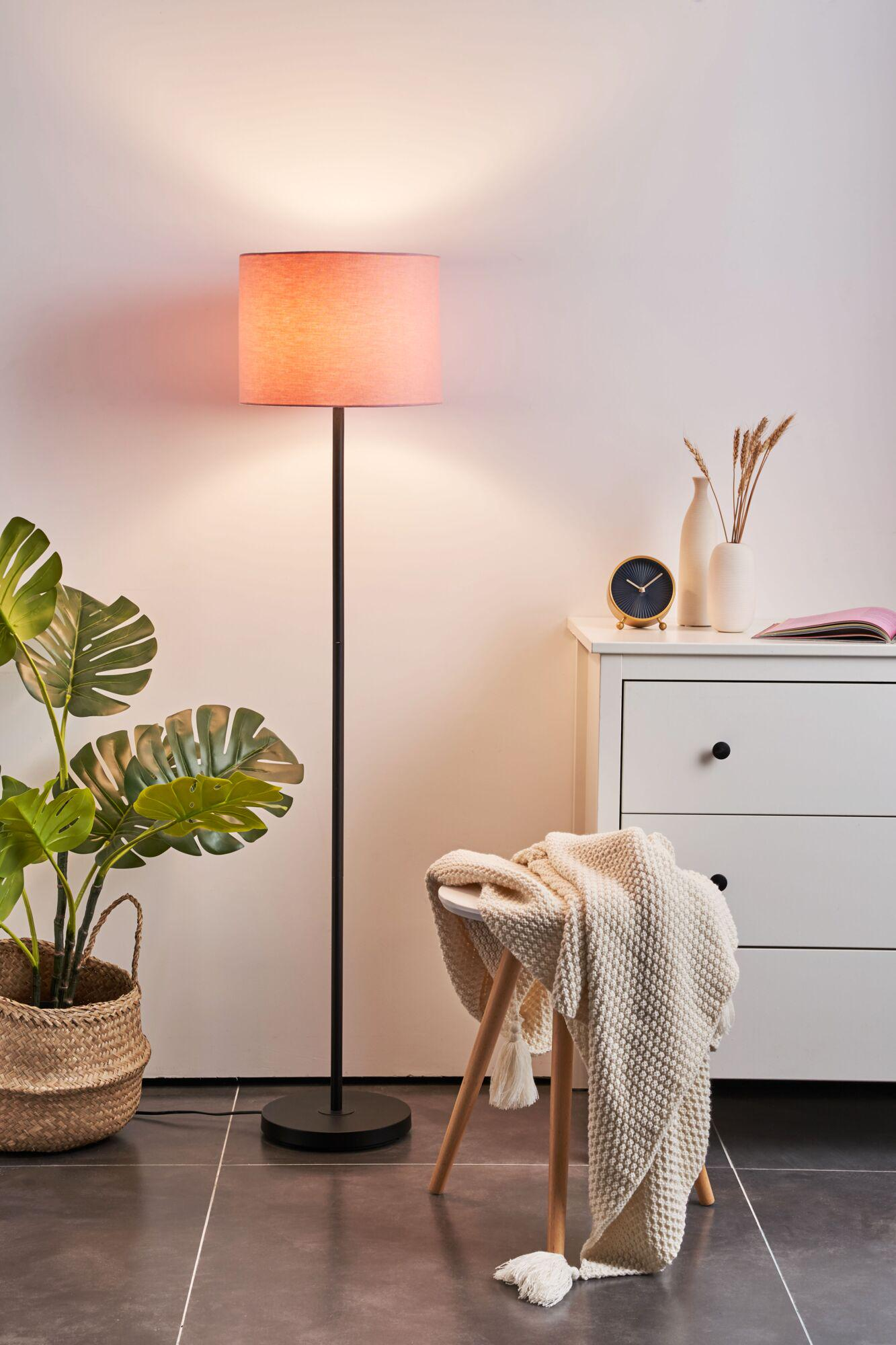 Lampa podłogowa rzuca ciepły blask na roślinę i białą komodę z dekoracjami.