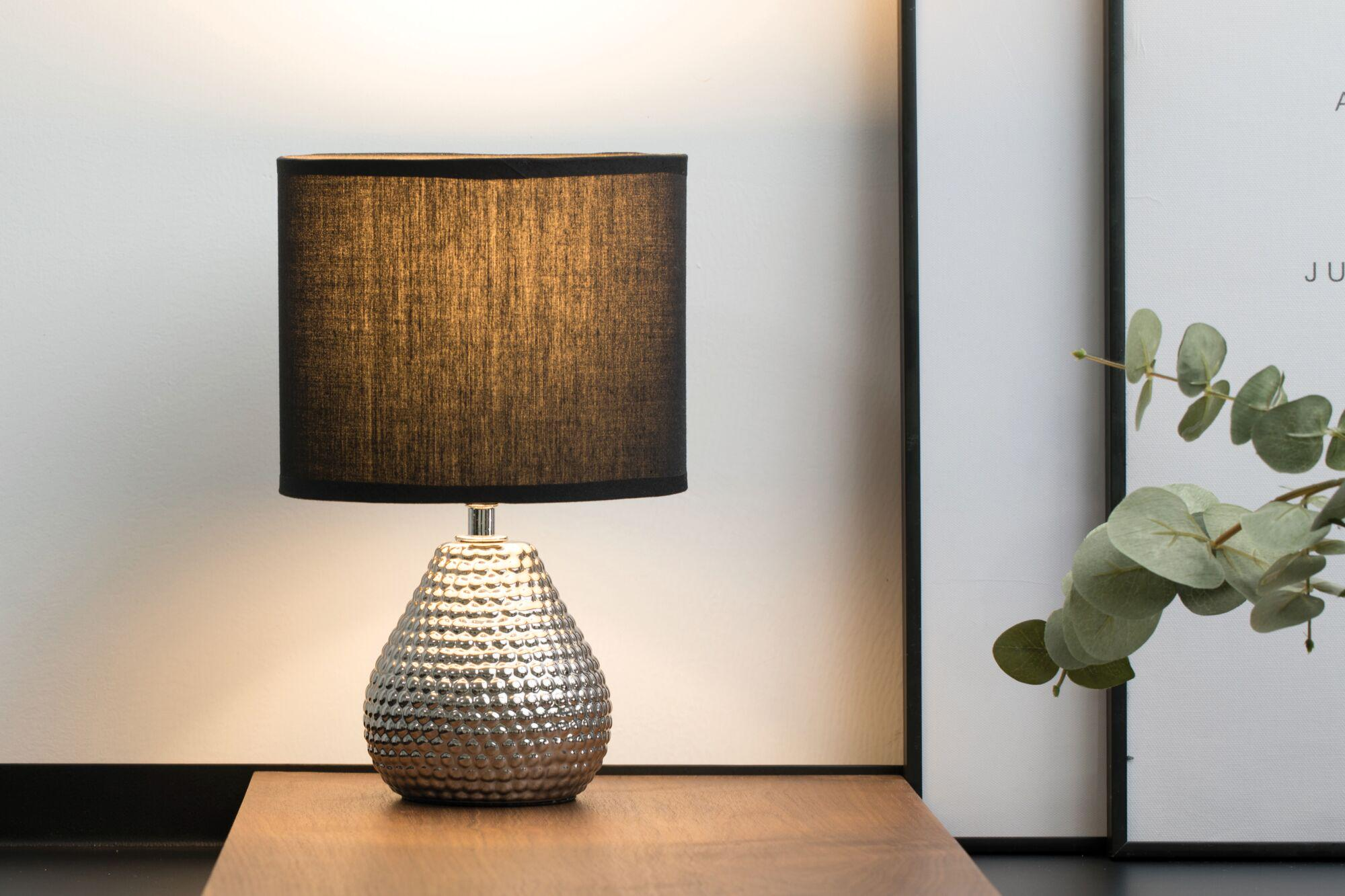 Srebrna lampa z czarnym kloszem stoi na drewnianym stole.