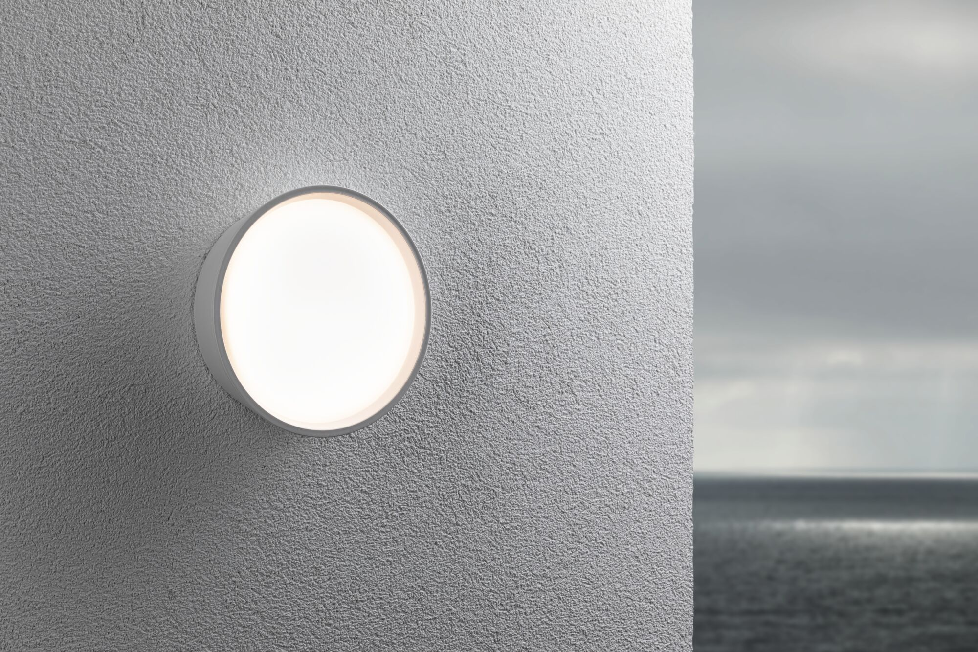 Eine runde Wandleuchte mit hellem Licht leuchtet auf einer strukturierten grauen Wand neben einem Meer.