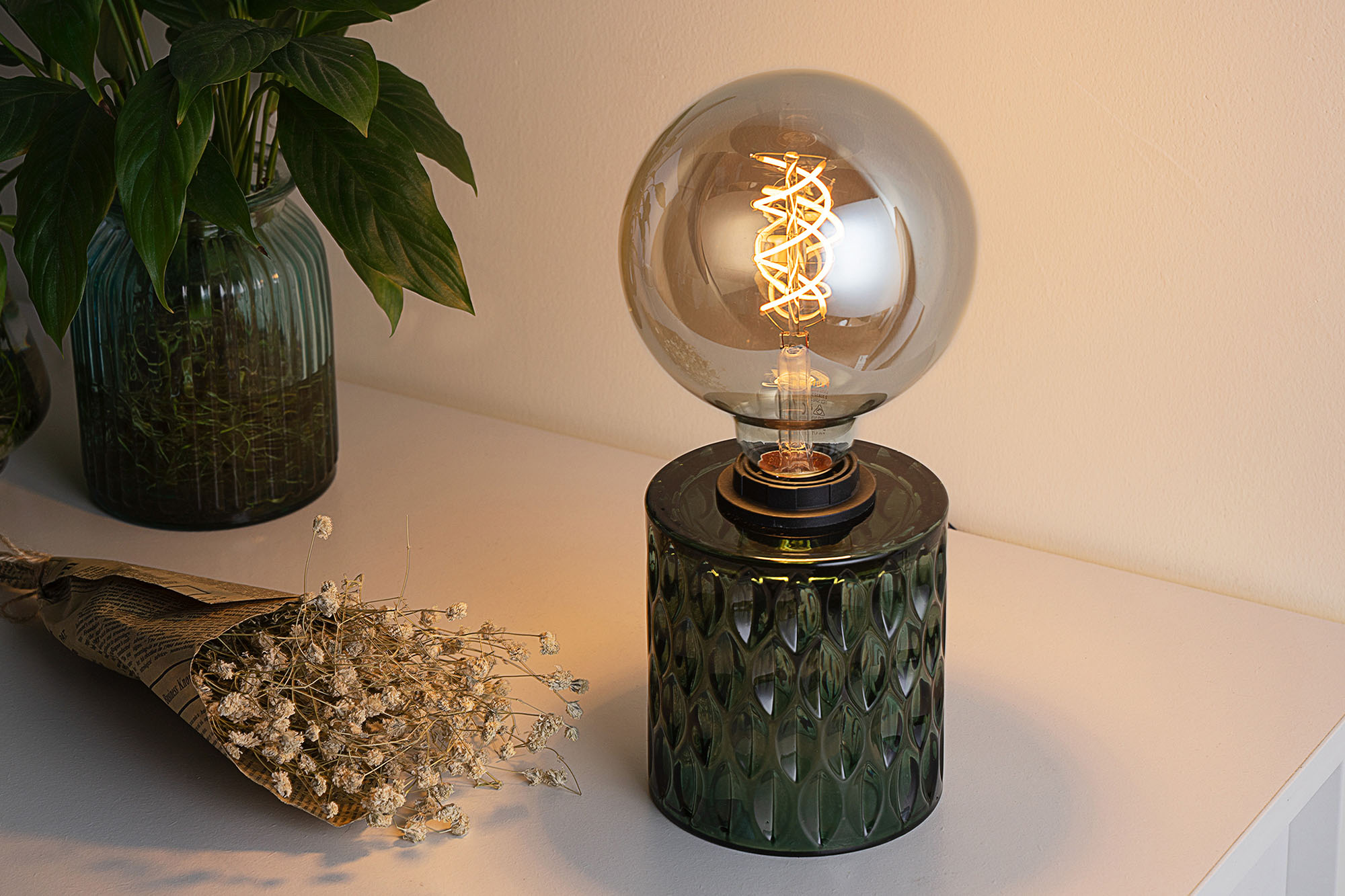 Lampa ze szklaną bańką i żarnikiem stoi obok rośliny i bukietu suszonych kwiatów.