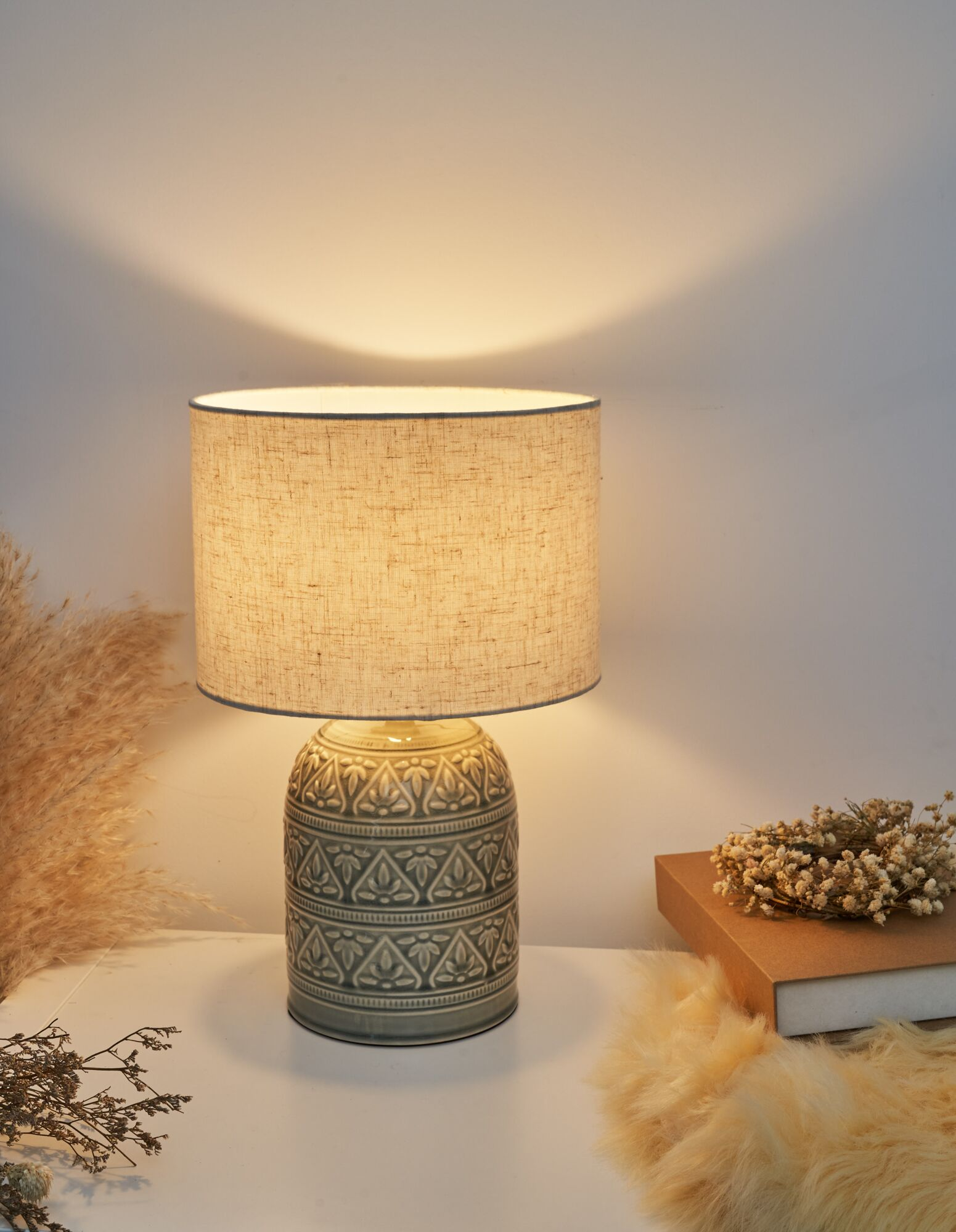 Wzorzysta lampa na stole, obok suszonych kwiatów i dywanika z owczej skóry.