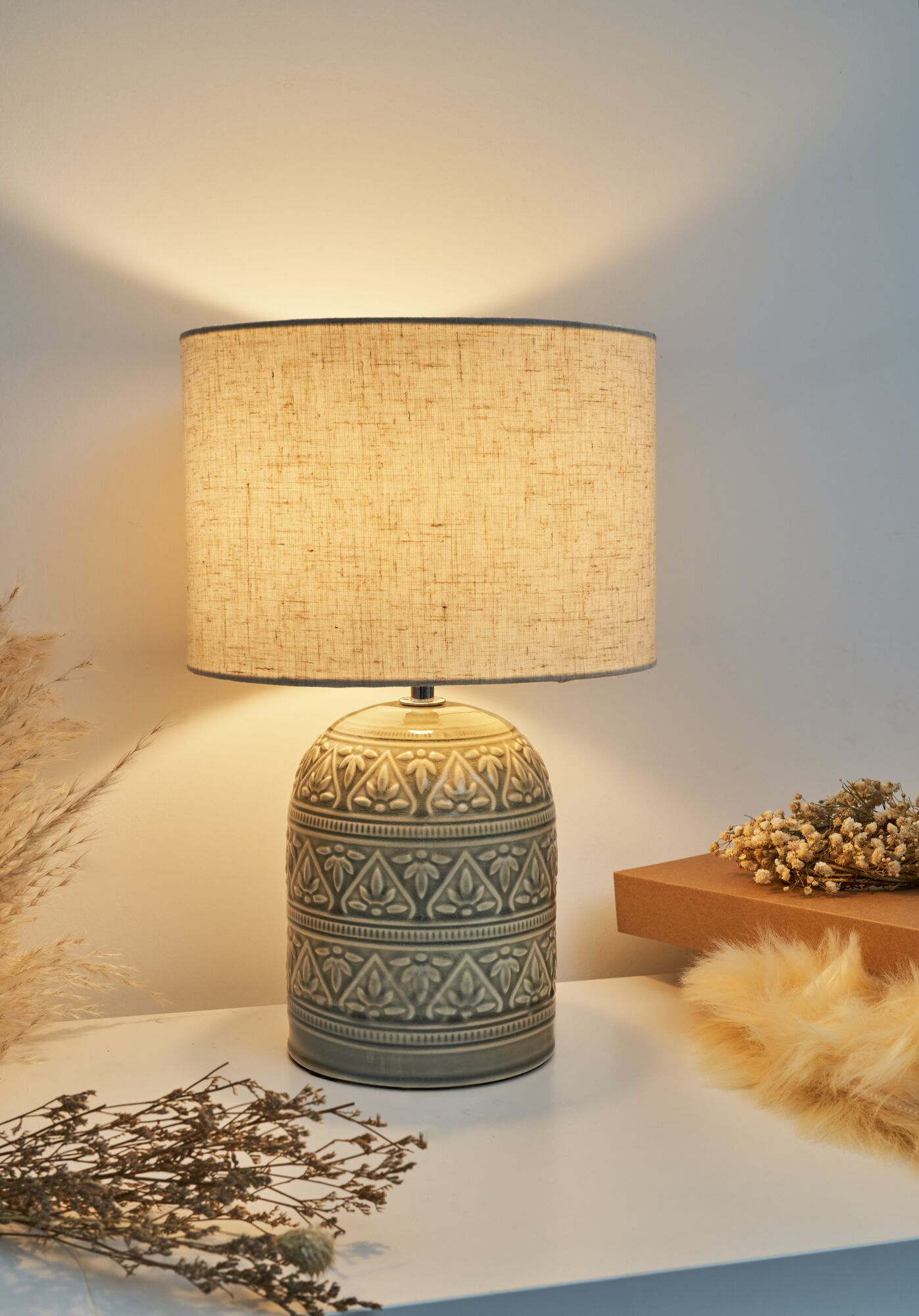 Szara ceramiczna lampa ze wzorzystą podstawą i lnianym kloszem stoi na białej powierzchni.