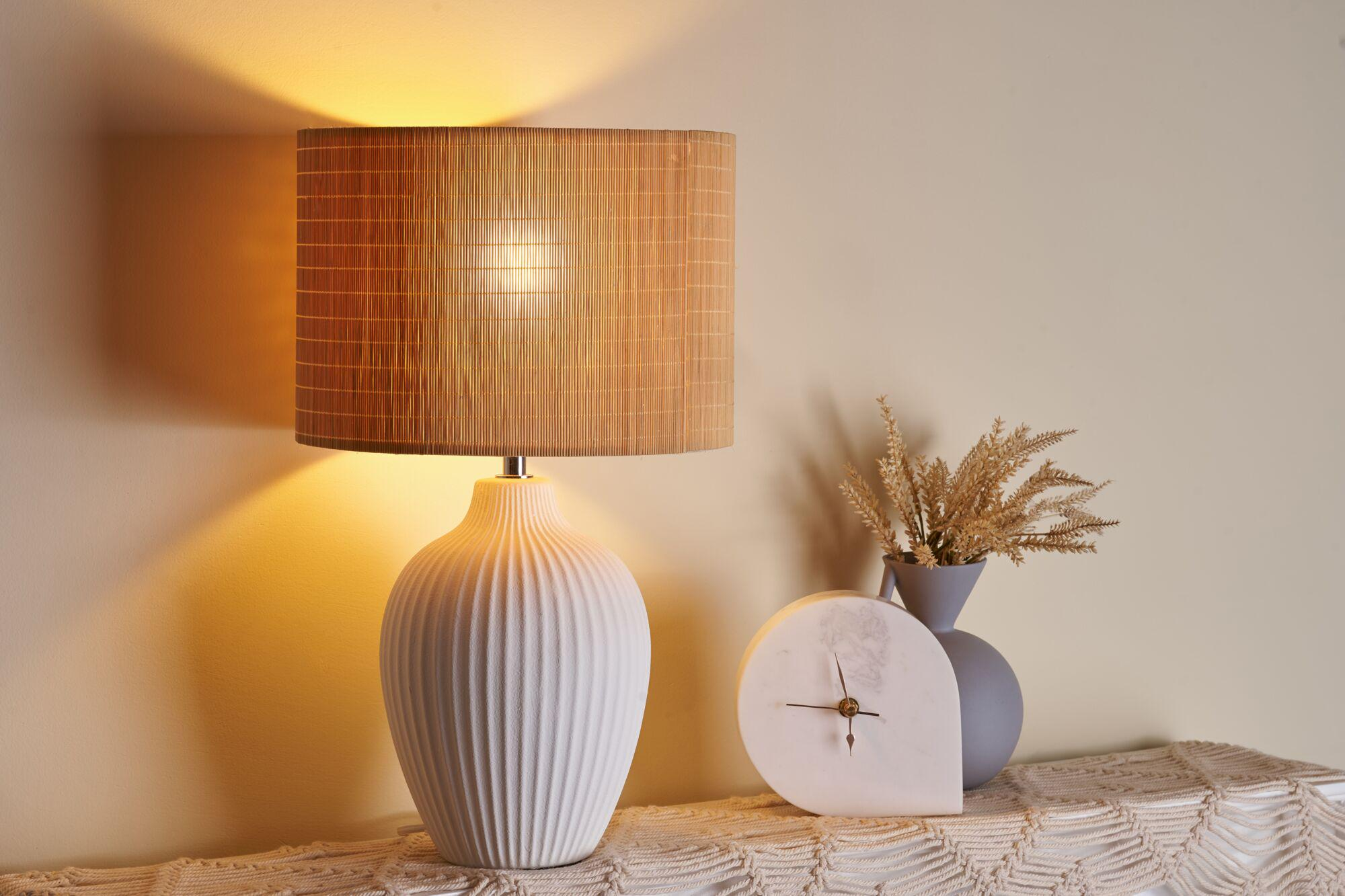 Biała lampa z bambusowym kloszem stoi obok szarego wazonu z suszonymi kwiatami i zegarem.