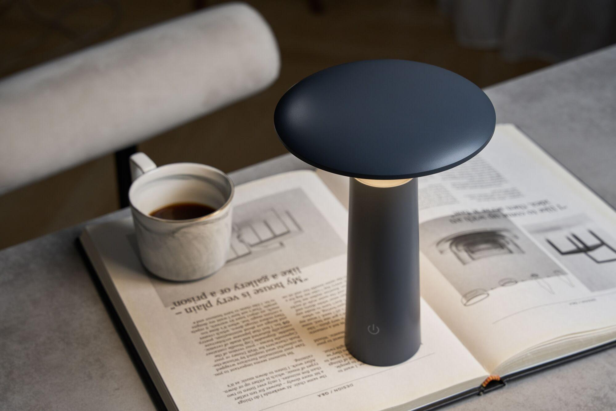 Ciemna lampka nocna stoi na otwartej książce obok filiżanki kawy.