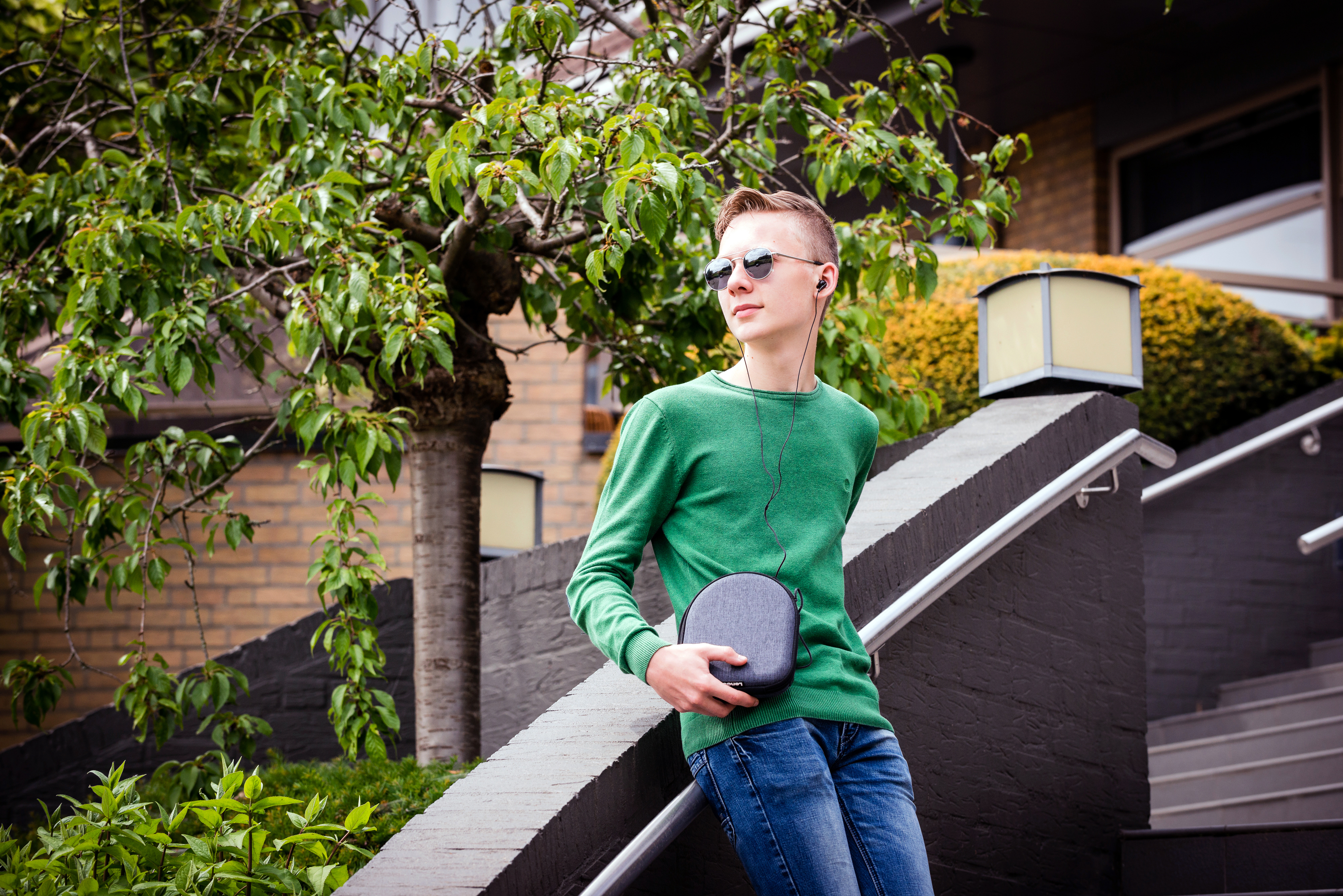 Un garçon en pull vert et jean avec écouteurs se tient dehors.