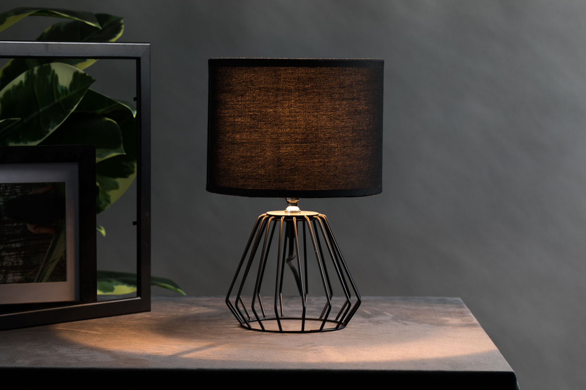 Czarna lampa stołowa z geometryczną drucianą podstawą stoi na stole.